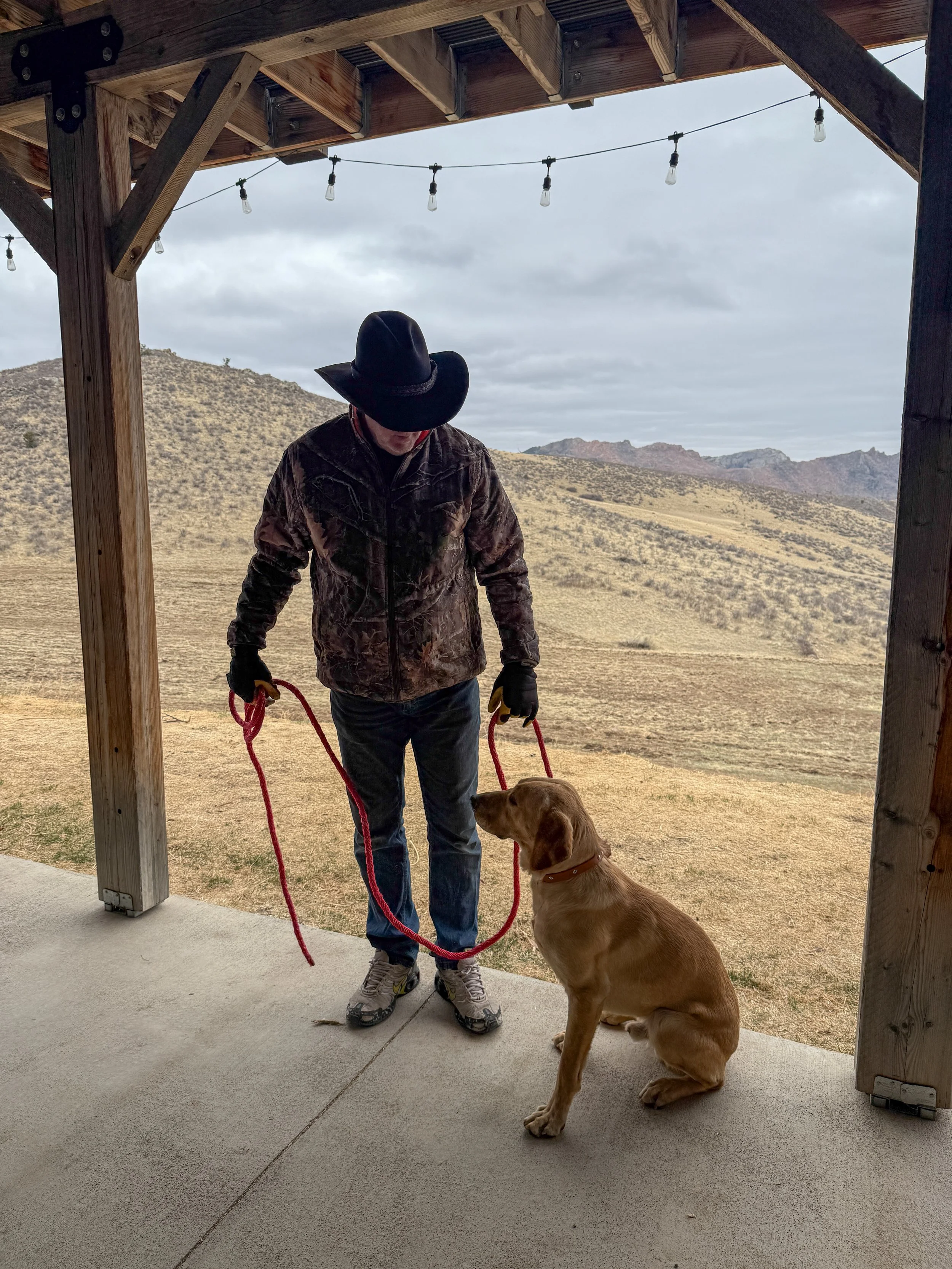 A man wearing a black cowboy hat, camouflage jacket, and jeans standing on a porch with a yellow Labrador retriever sitting in front of him. The man is holding a red leash attached to the dog's collar. The background shows a hilly landscape and cloud