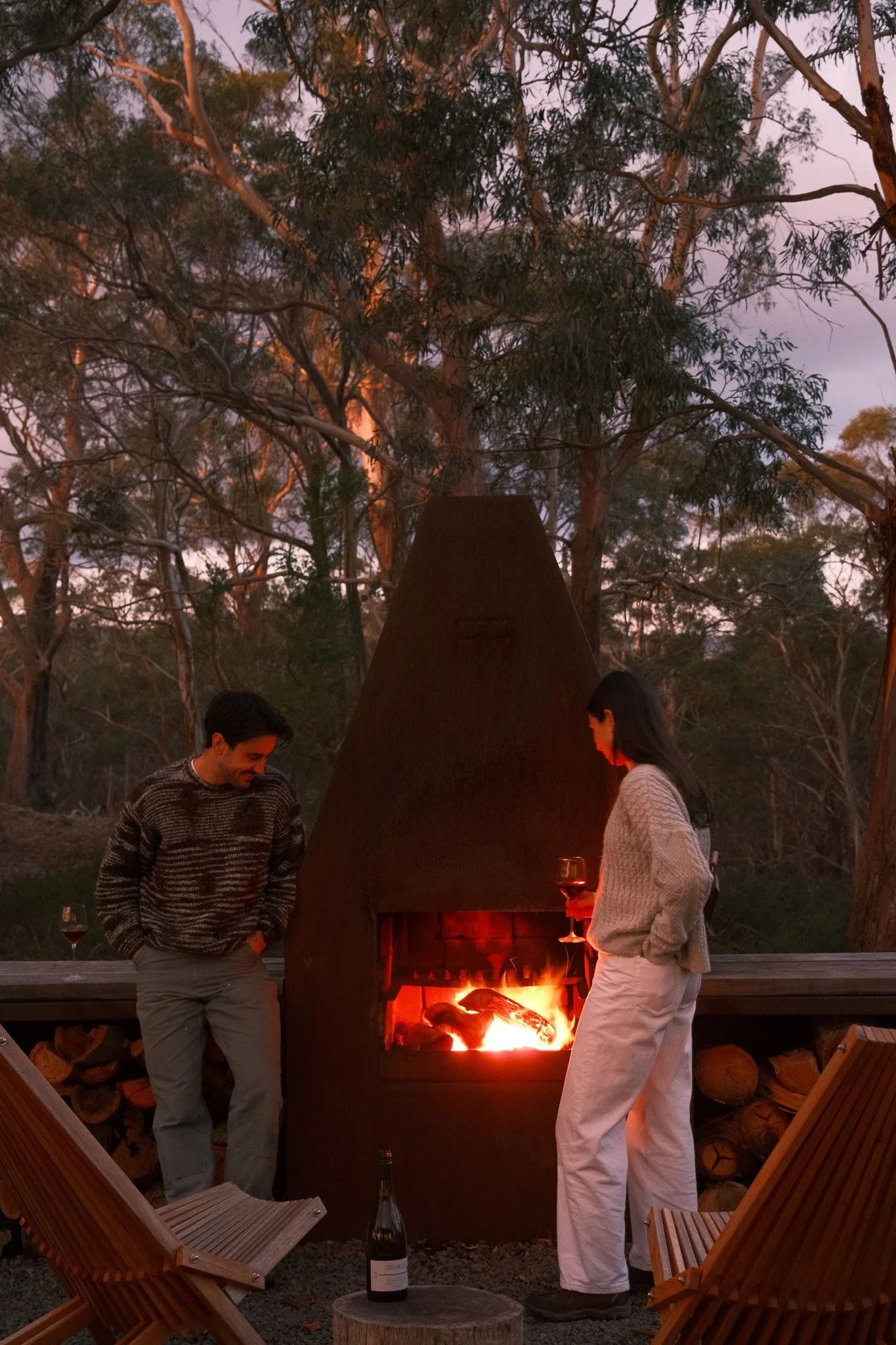 A man and woman stand near an outdoor fireplace with a fire burning, surrounded by trees at dusk or sunset. The woman holds a glass of red wine, and a bottle of wine is on a small wooden table nearby, with two wooden chairs in the foreground.