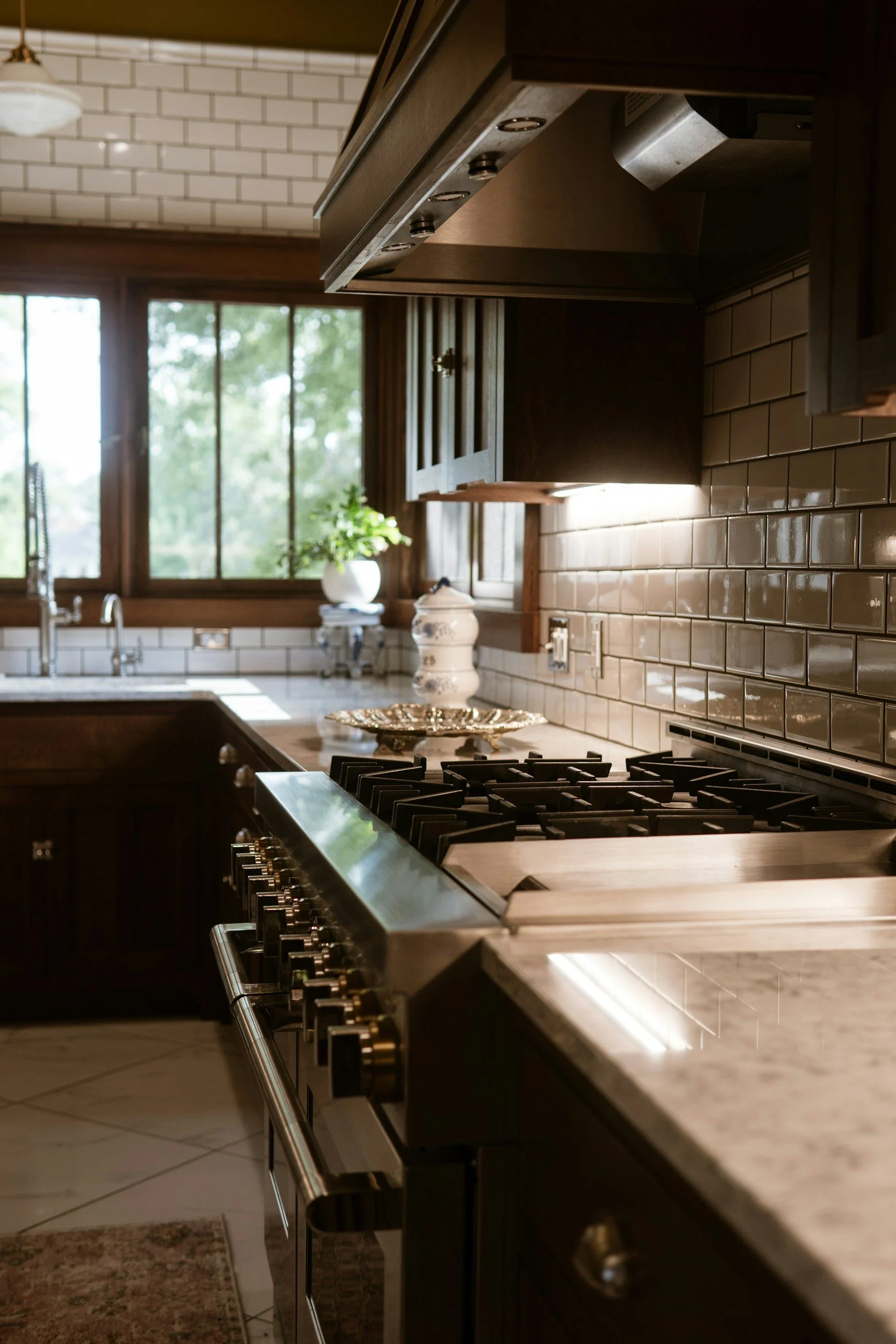A modern kitchen with dark wooden cabinets, a stainless steel gas stove, a white tiled backsplash, and a window with a view of trees outside.
