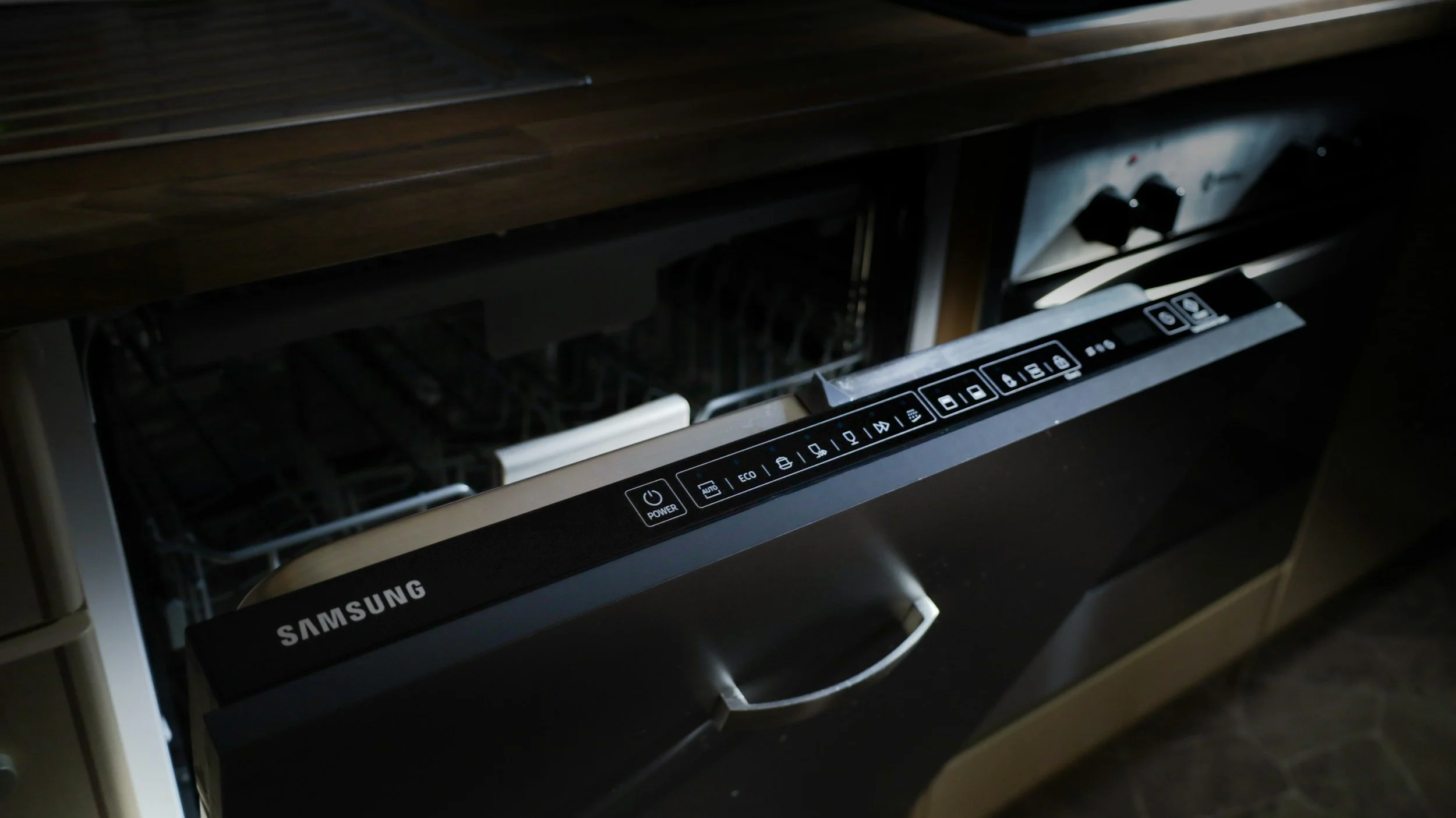 Samsung dishwasher with open door showing control panel and interior racks in a dark kitchen.