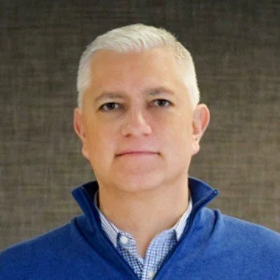 A middle-aged man with short, white hair wearing a blue jacket over a collared shirt, standing against a dark, textured background.