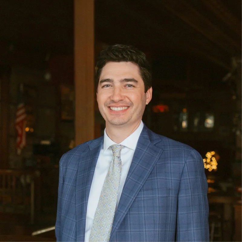A smiling man in a blue plaid suit with a white shirt and a patterned tie, standing indoors with a warm, rustic background.