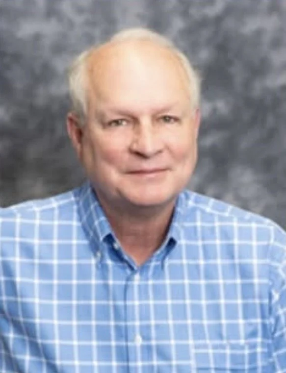 A middle-aged man with fair skin and short, light-colored hair, wearing a blue and white checkered shirt, posing in front of a gray textured background.