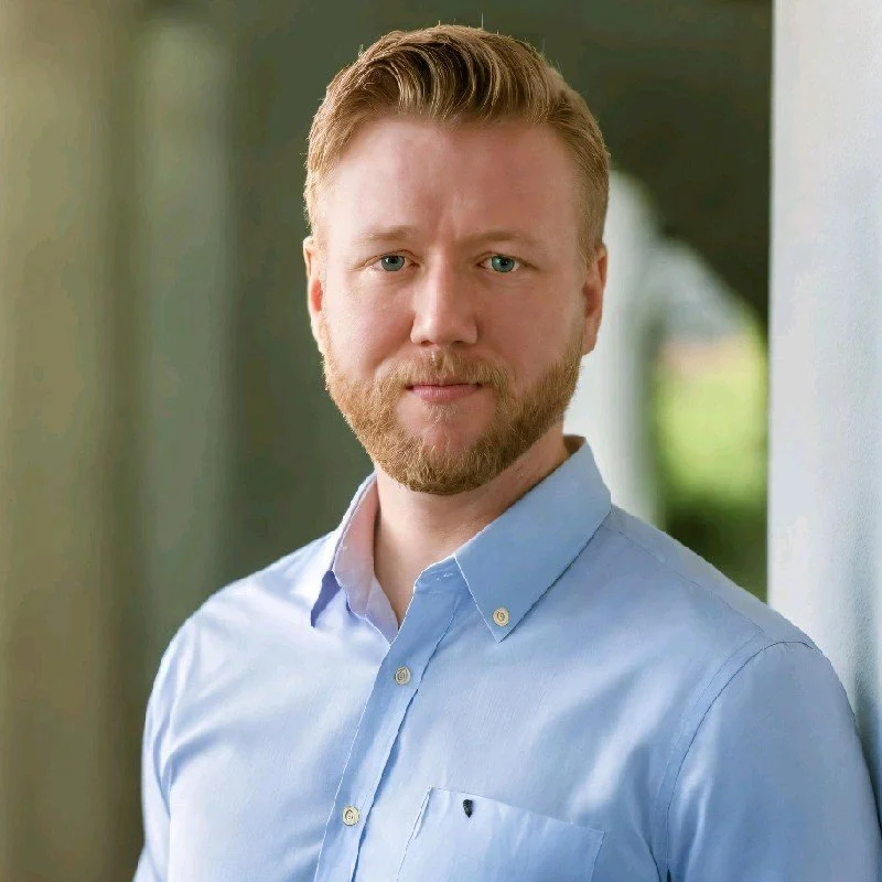 A man with red hair and a beard wearing a light blue button-up shirt, standing outdoors beside a white wall with greenery in the background.