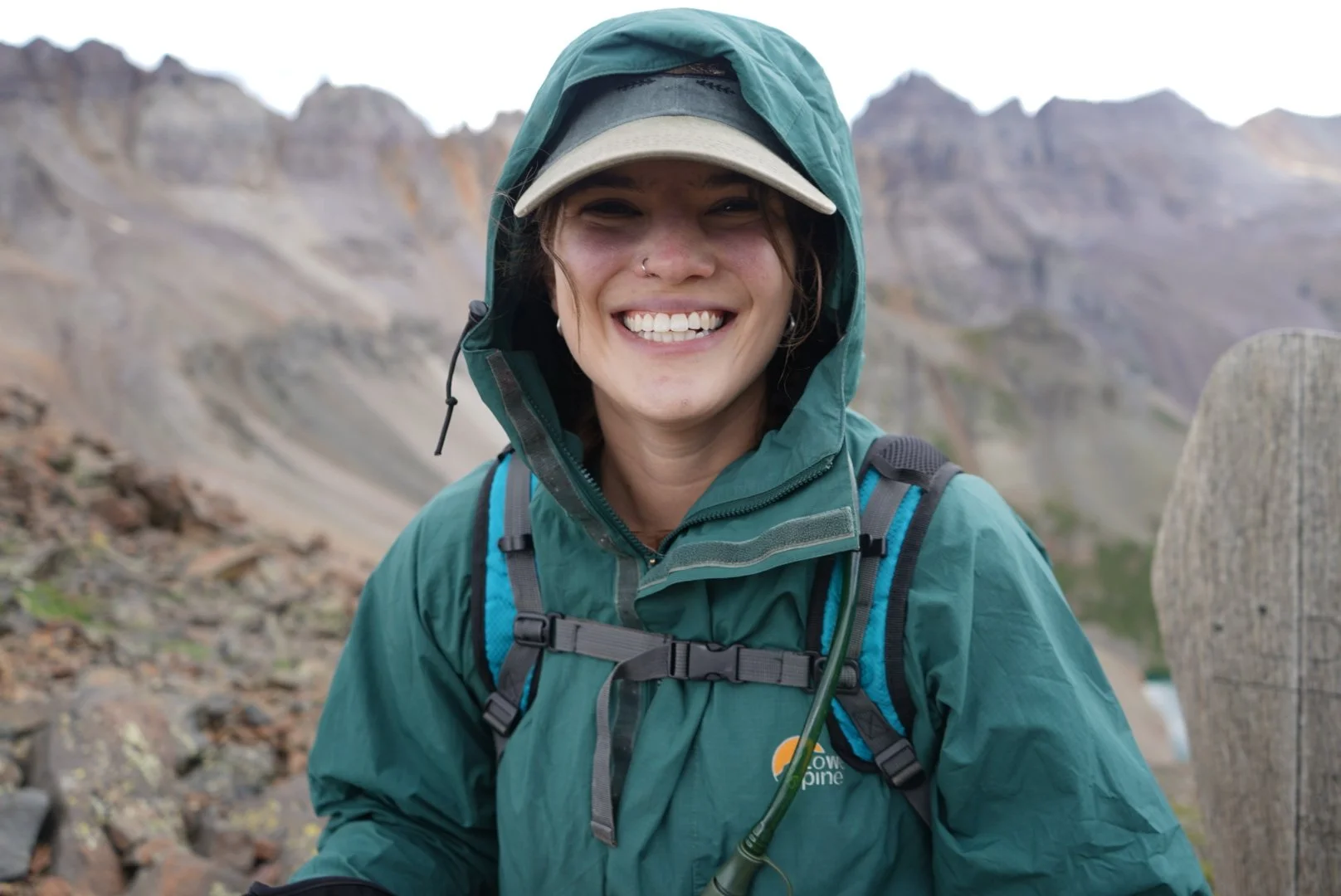 Smiling woman wearing outdoor gear and hoodie on mountain trail with rocky peaks in background.