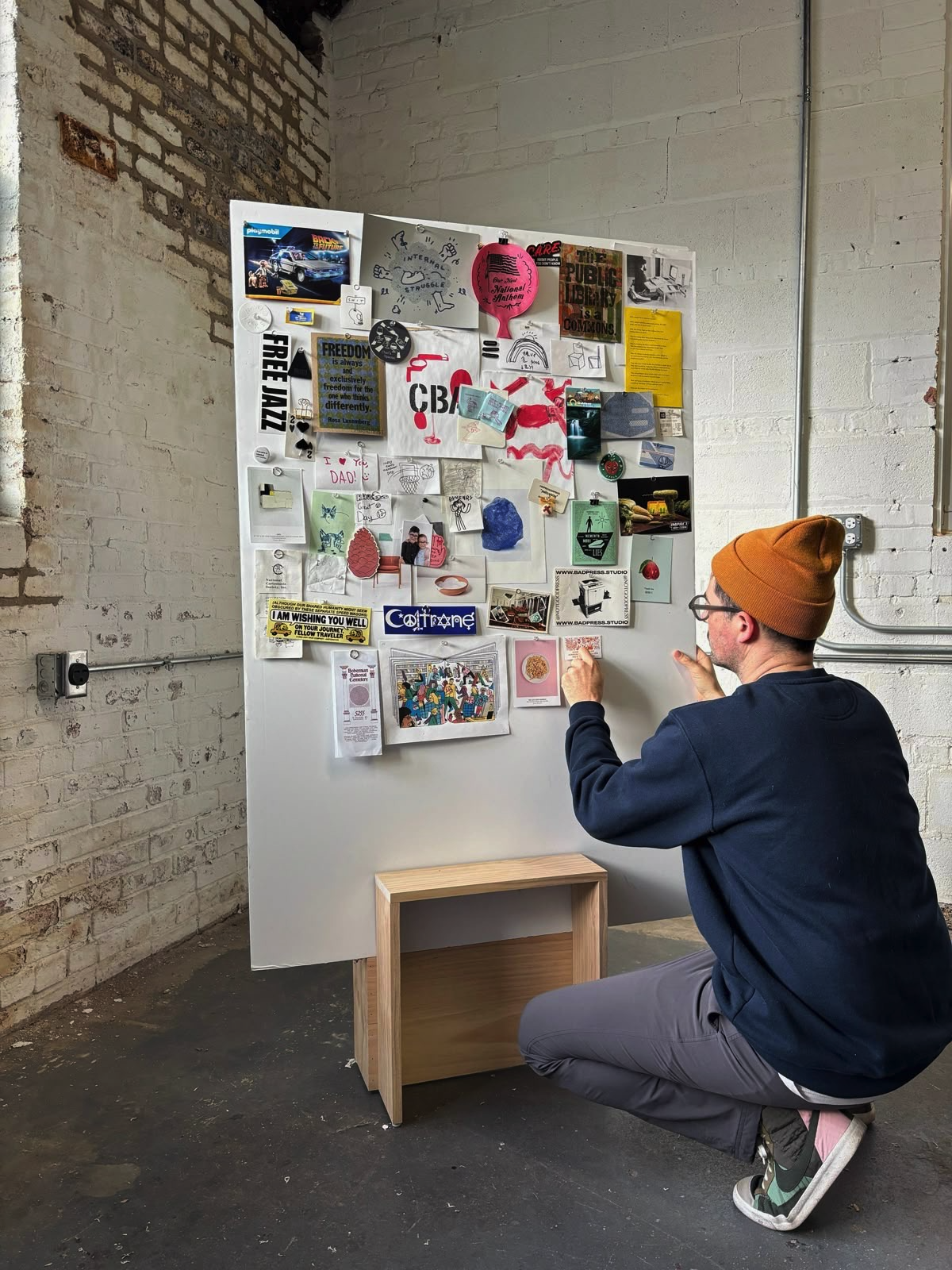 A man wearing a brown beanie, glasses, a navy sweatshirt, gray pants, and sneakers is crouching in front of a white bulletin board filled with various photos, notes, and artwork. The bulletin board is against a white brick wall with some exposed brick and an electrical outlet visible.