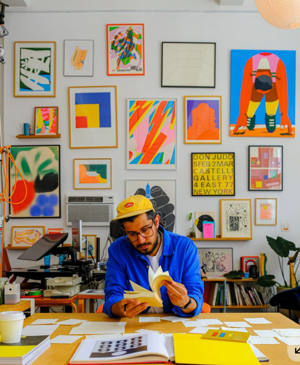 Man sitting at a desk reading a book in an art studio or gallery with colorful abstract artwork displayed on the wall behind him.