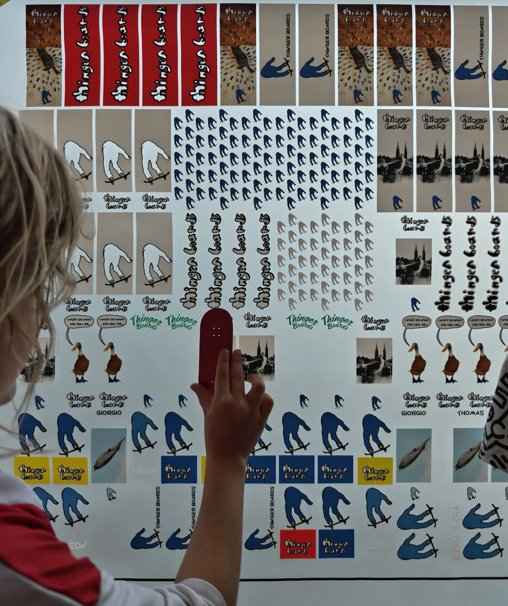 A person pointing at a series of stickers on a wall, including skateboarder and duck illustrations, with the words "Thinger Boards" and "Giorgio" visible on some stickers.