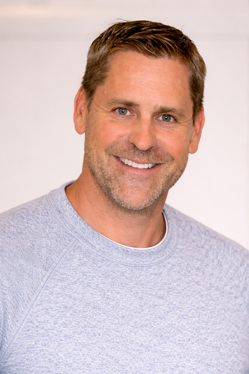 A smiling middle-aged man with short brown hair, blue eyes, and a light beard, wearing a light gray sweatshirt, standing against a plain background.