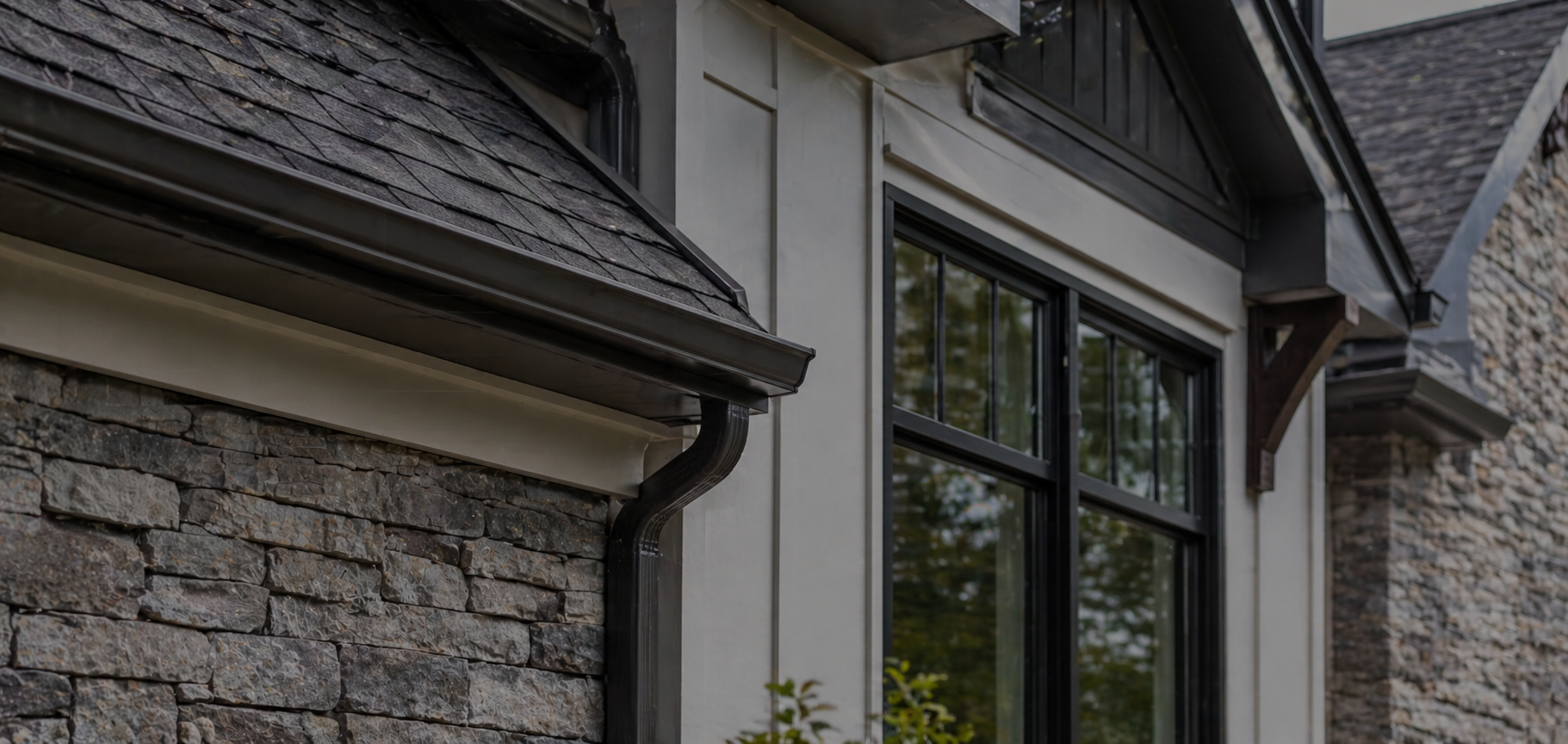 Close-up of the corner of a house showing stone siding, a window with a black frame, and a black gutter system with a downspout.