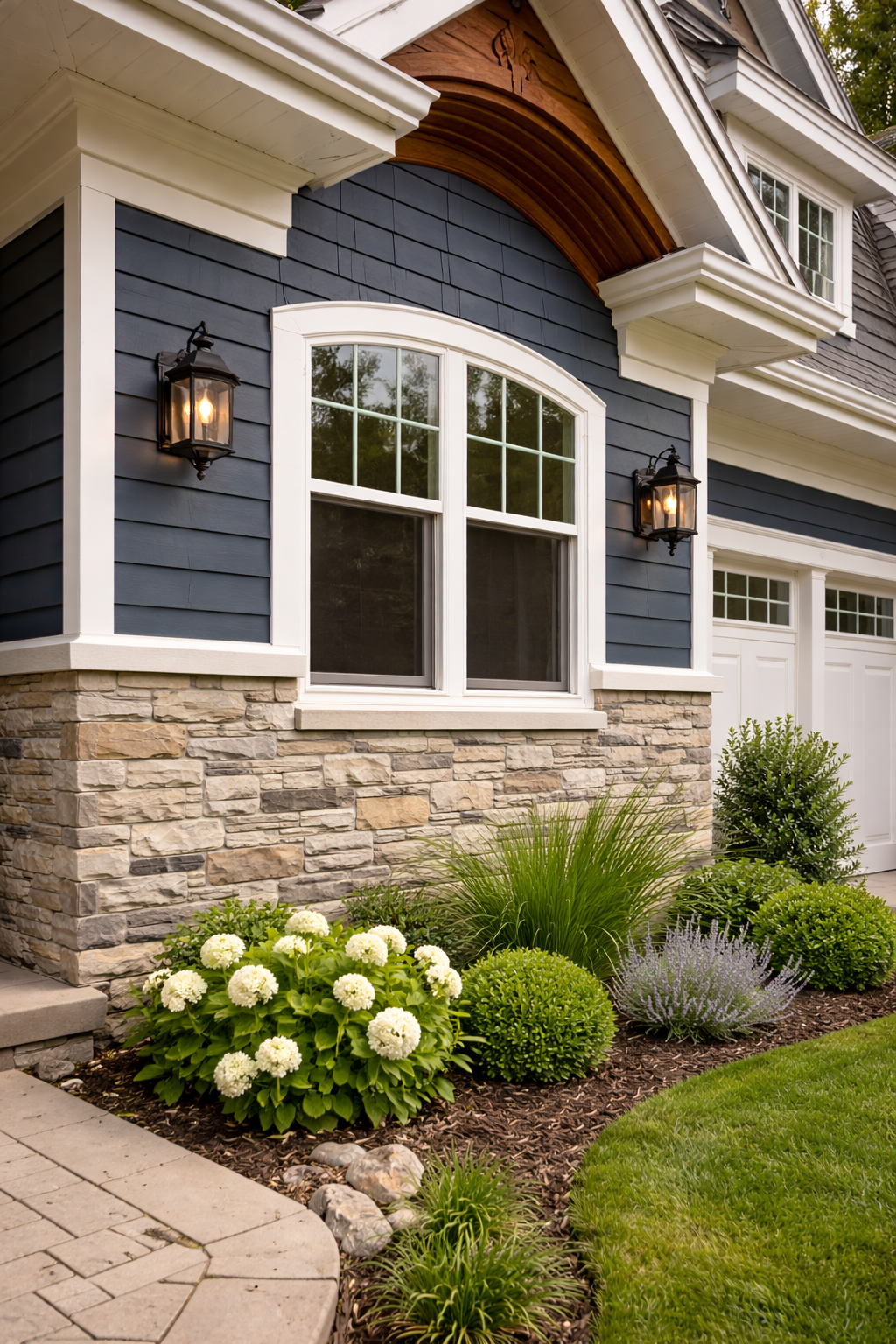 Close-up of the exterior of a house with dark blue siding, white trim, stone foundation, large window, and decorative arched wood accent above the window. Two wall-mounted lamps are on either side of the window. There is a landscaped garden with white hydrangeas, green shrubs, and ornamental grasses in front of the house, with a paved walkway nearby.