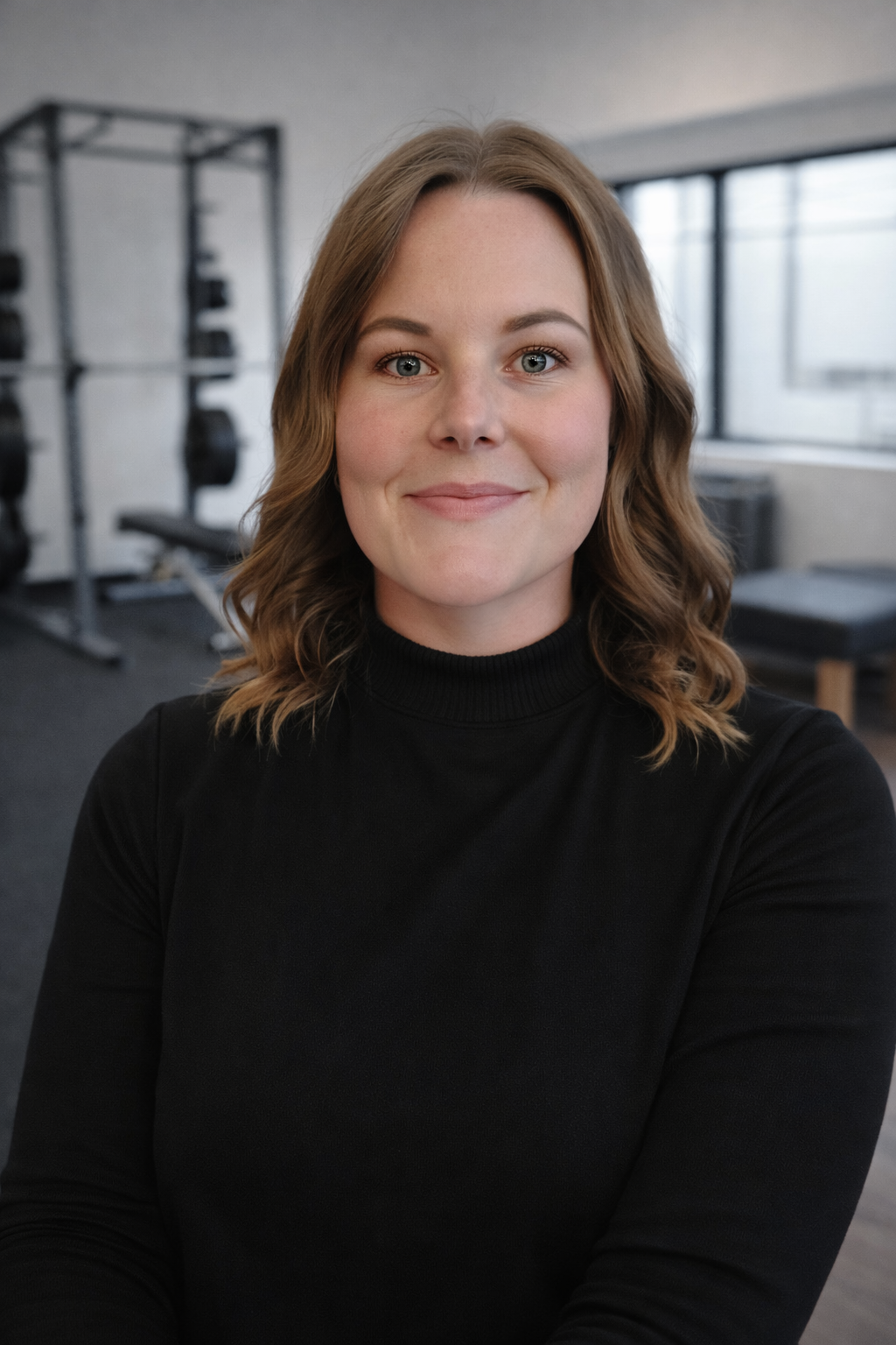A woman with shoulder-length wavy light brown hair, blue eyes, and a friendly smile, wearing a black turtleneck, standing inside a gym with workout equipment in the background.