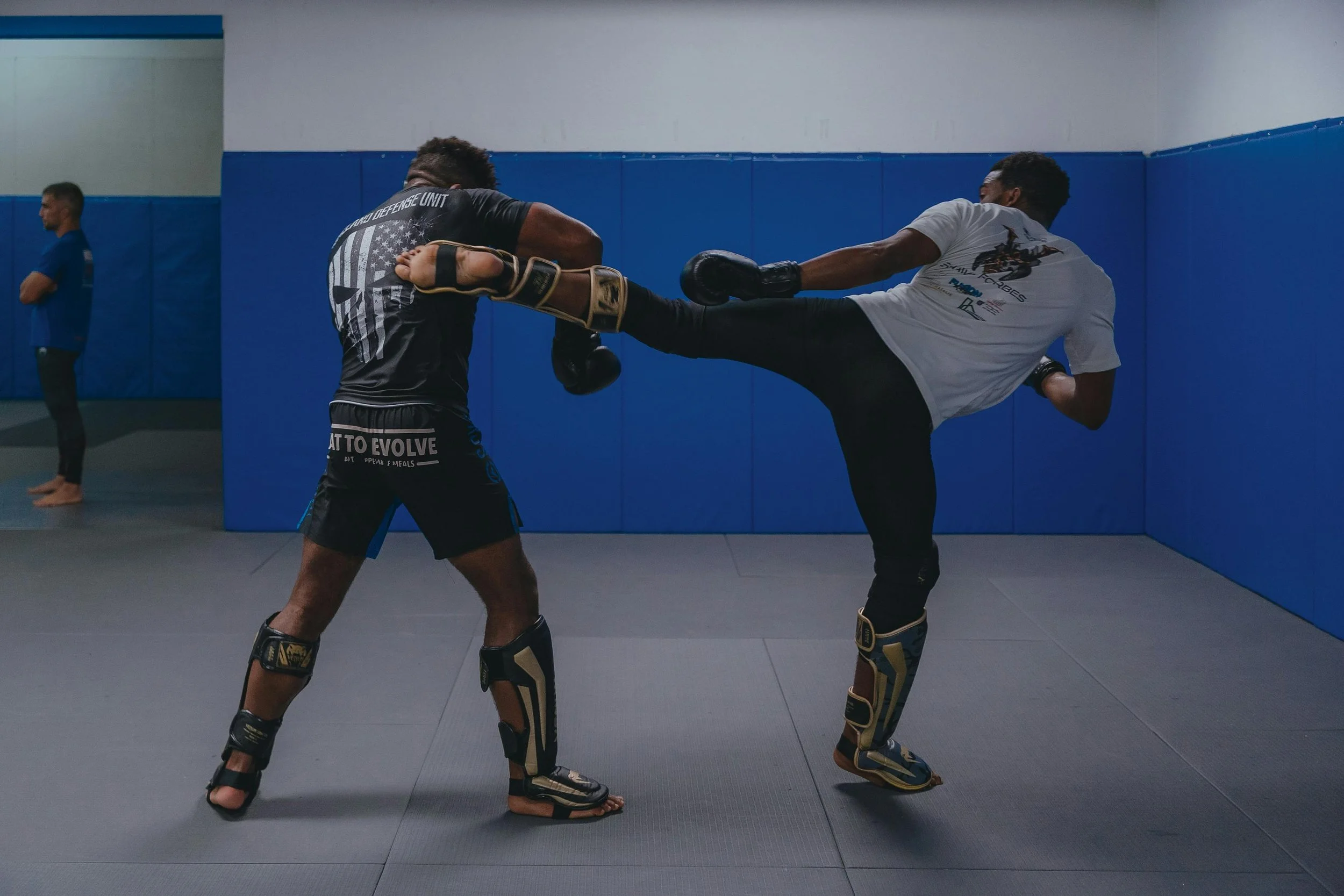 Two men practicing martial arts in a gym. One man is delivering a high kick towards the other's face, who is blocking with his arm. They are wearing protective gear including knee and shin pads.