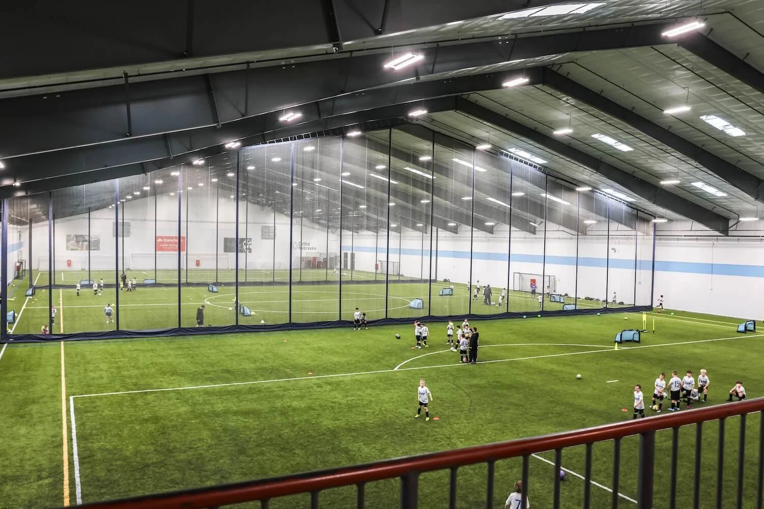 Indoor soccer field with young players practicing. The field is covered with artificial turf and is enclosed by a large mesh net. Children are seen engaging in drills and activities, with training equipment scattered around.