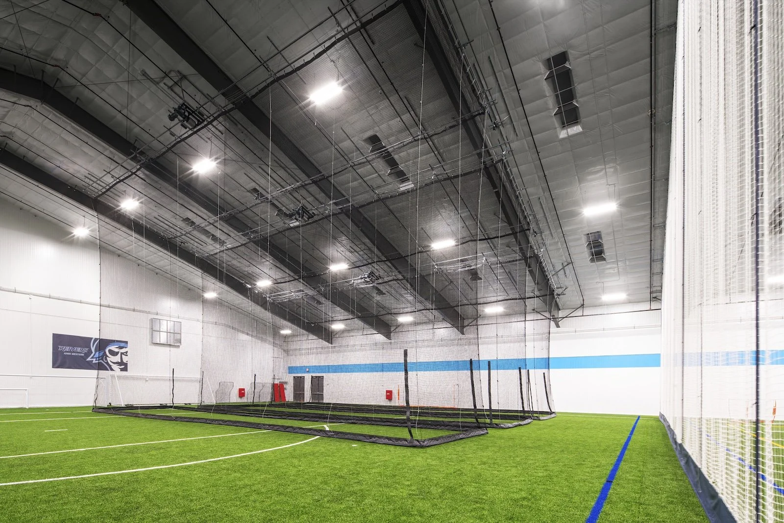 Indoor sports training facility with artificial turf field, batting cage with nets, and overhead lighting.
