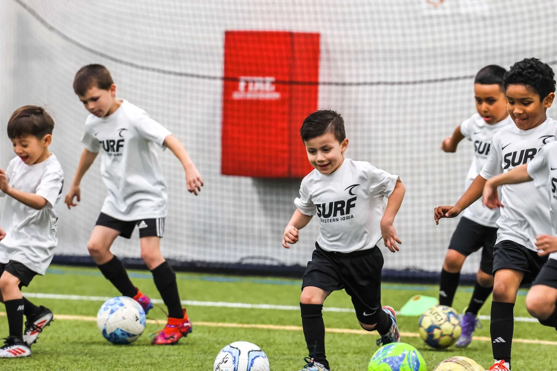 Young boys playing soccer indoors on a green turf field, wearing white jerseys with 'SURF' logo, black shorts, and soccer shoes, with colorful soccer balls in front of them.