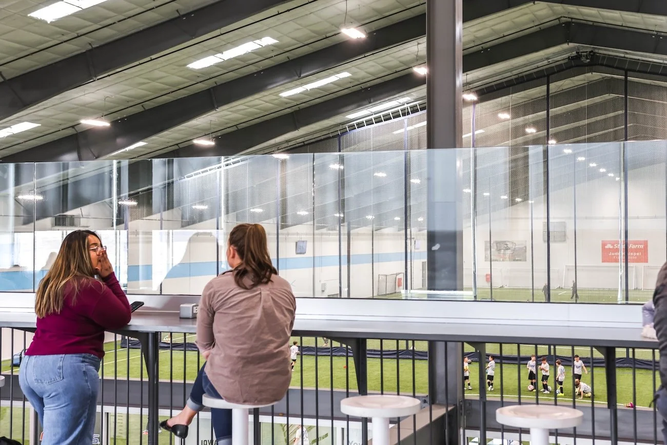 Two women talking at a sports bar with an indoor soccer field and players visible through a glass partition behind them.