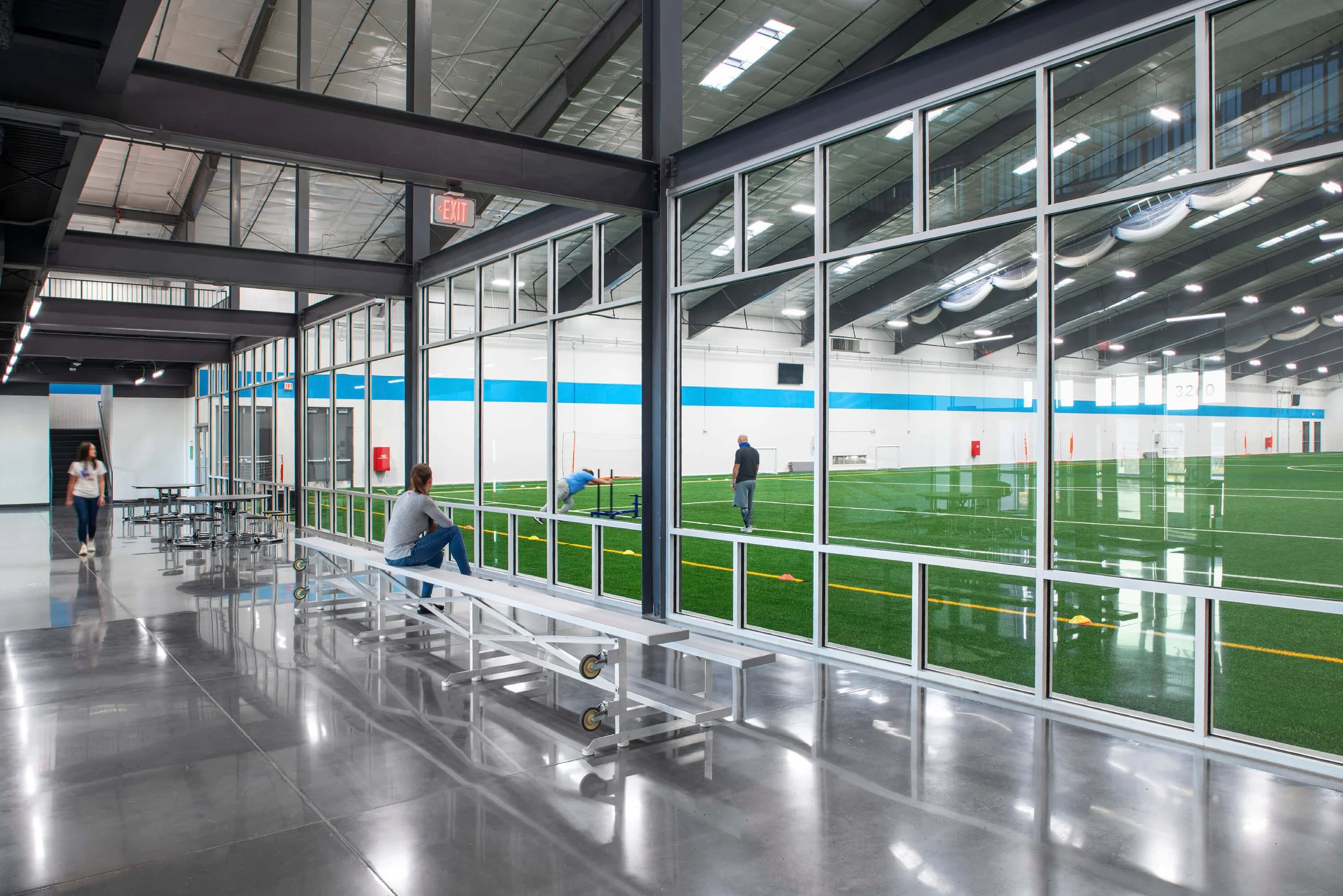 Interior of an indoor sports facility with a large glass window displaying a green turf field where people are practicing football drills; inside are benches and a woman sitting, with two women walking near the airlock entrance.
