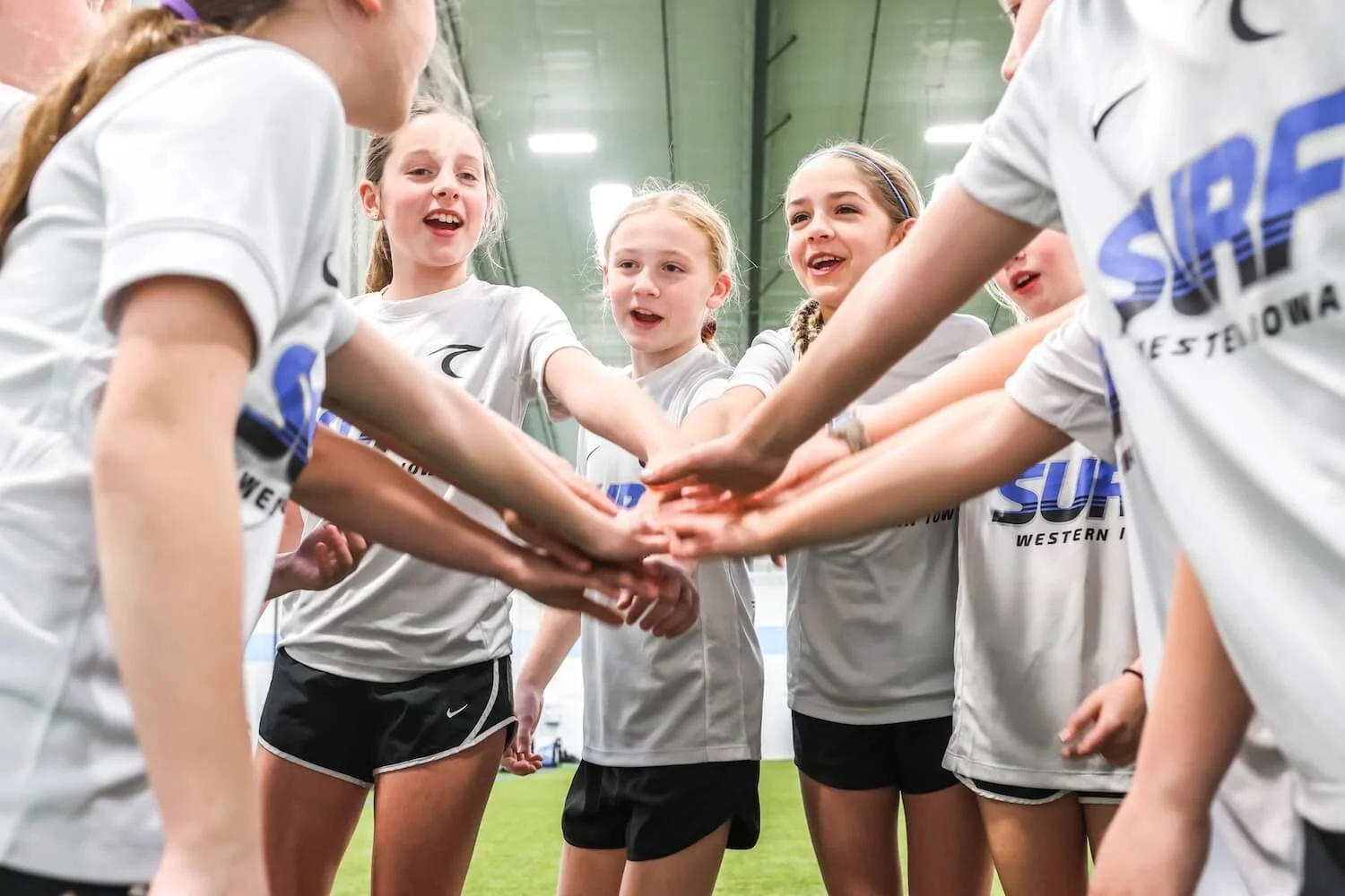 A group of young female soccer players in white jerseys and black shorts huddled together inside a sports arena, placing their hands together in the center.