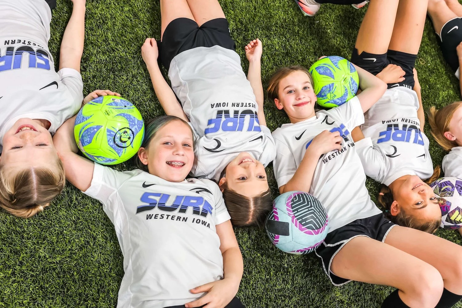 Children lying on grass in soccer uniforms, smiling, with soccer balls
