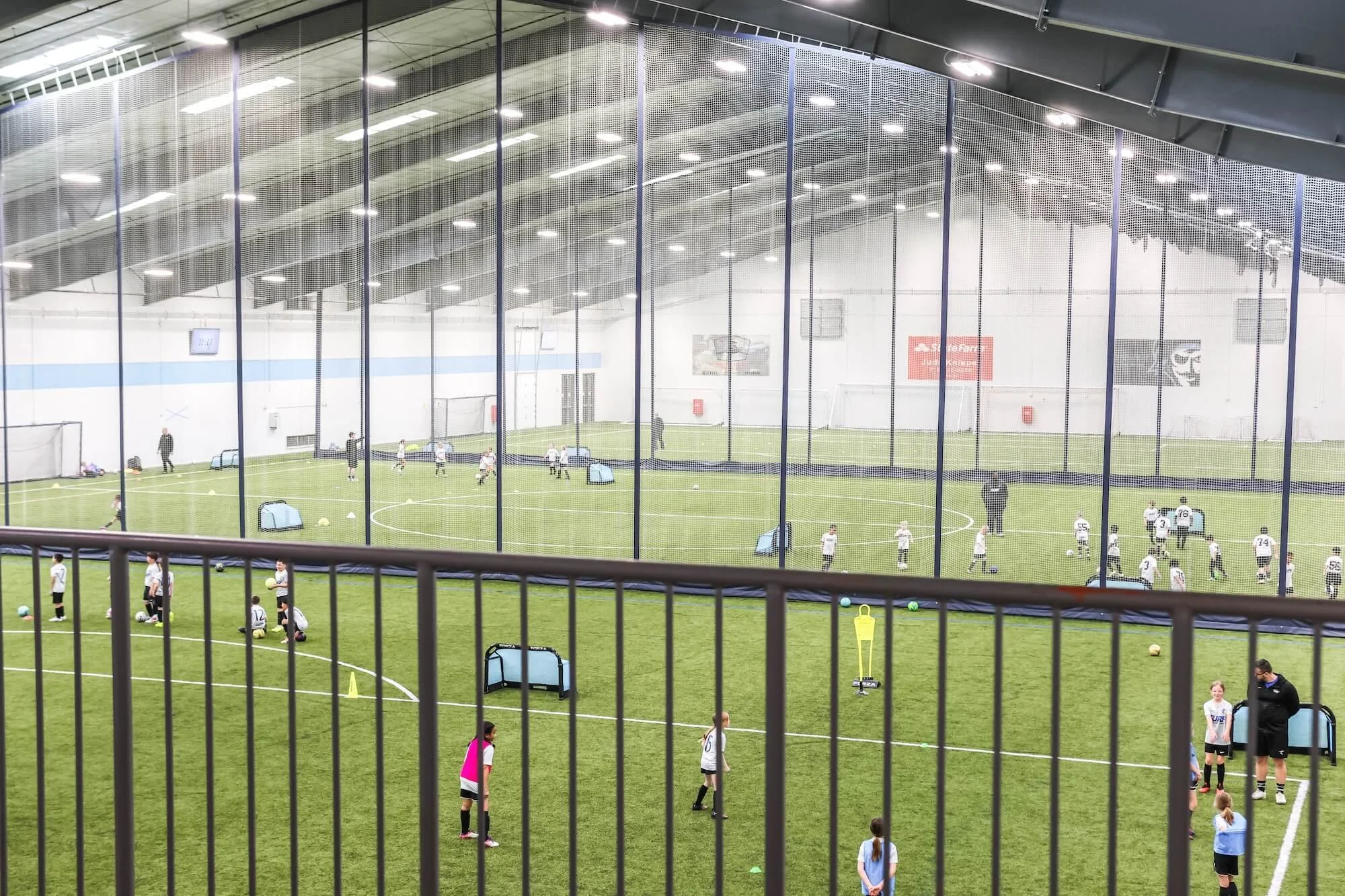 Indoor soccer training session with young players practicing on a green artificial turf field, separated by a net barrier, with coaches and training equipment visible.