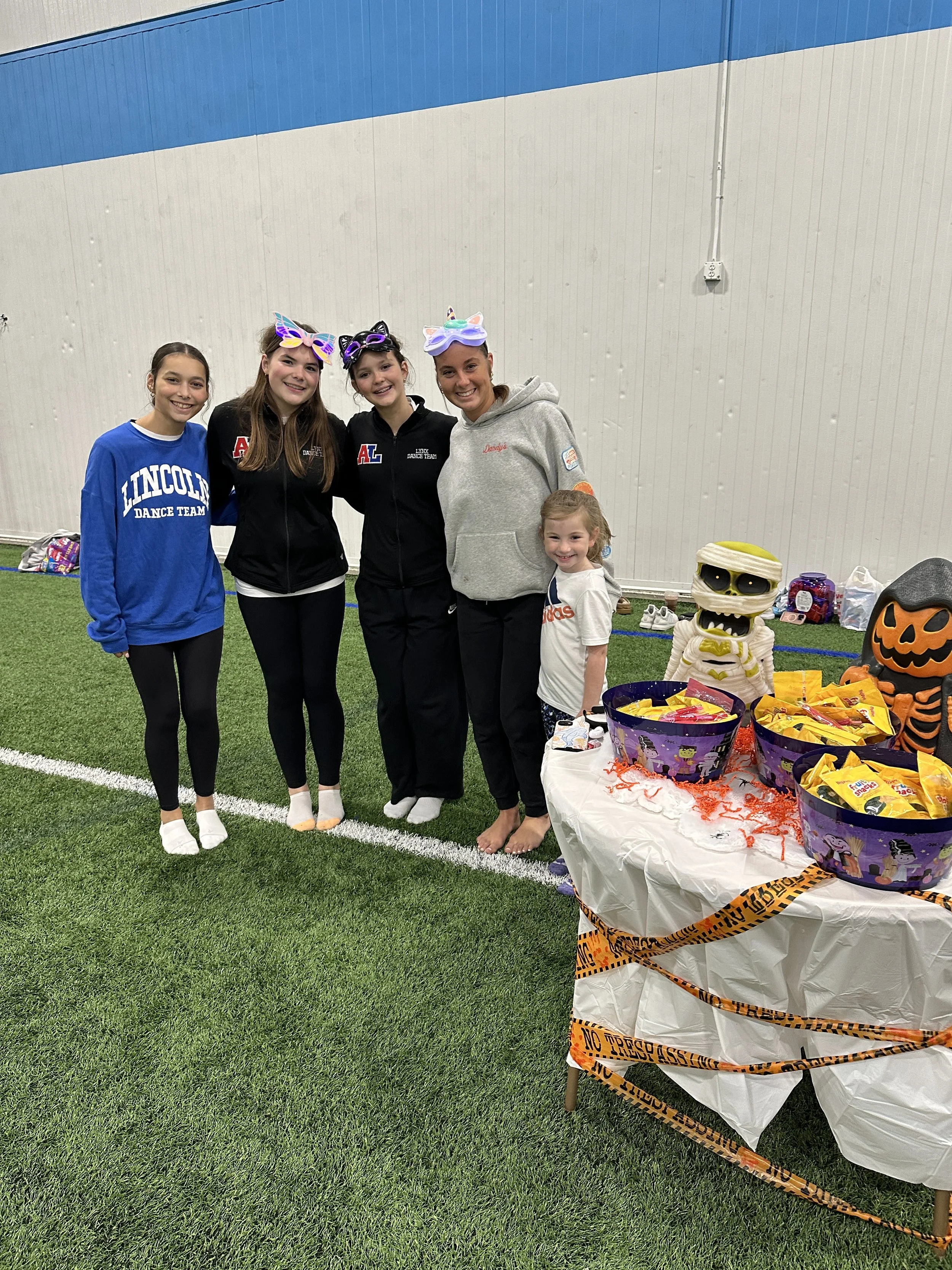 Group of four women and one young girl standing on an indoor turf field, dressed in casual sportswear, with Halloween-themed decorations and treats on a table nearby.