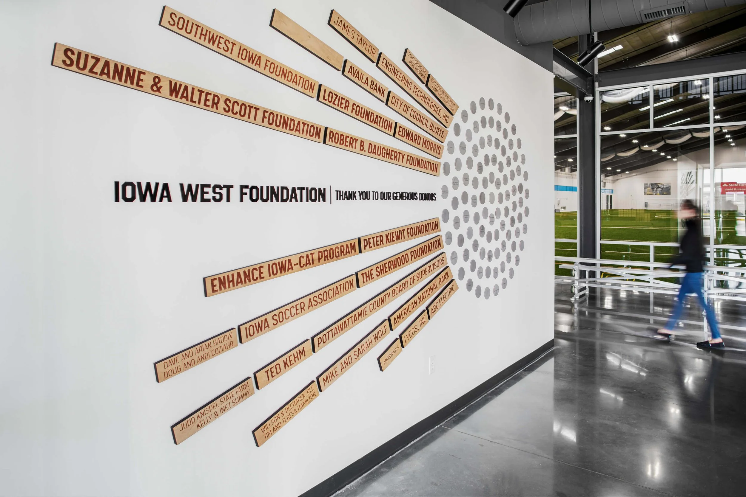 Wall display with wooden plaques listing donors for the Iowa West Foundation, including foundations, programs, and individuals, with a large circular ventilation or decorative element on the wall, and a person walking in a sports facility visible through windows.