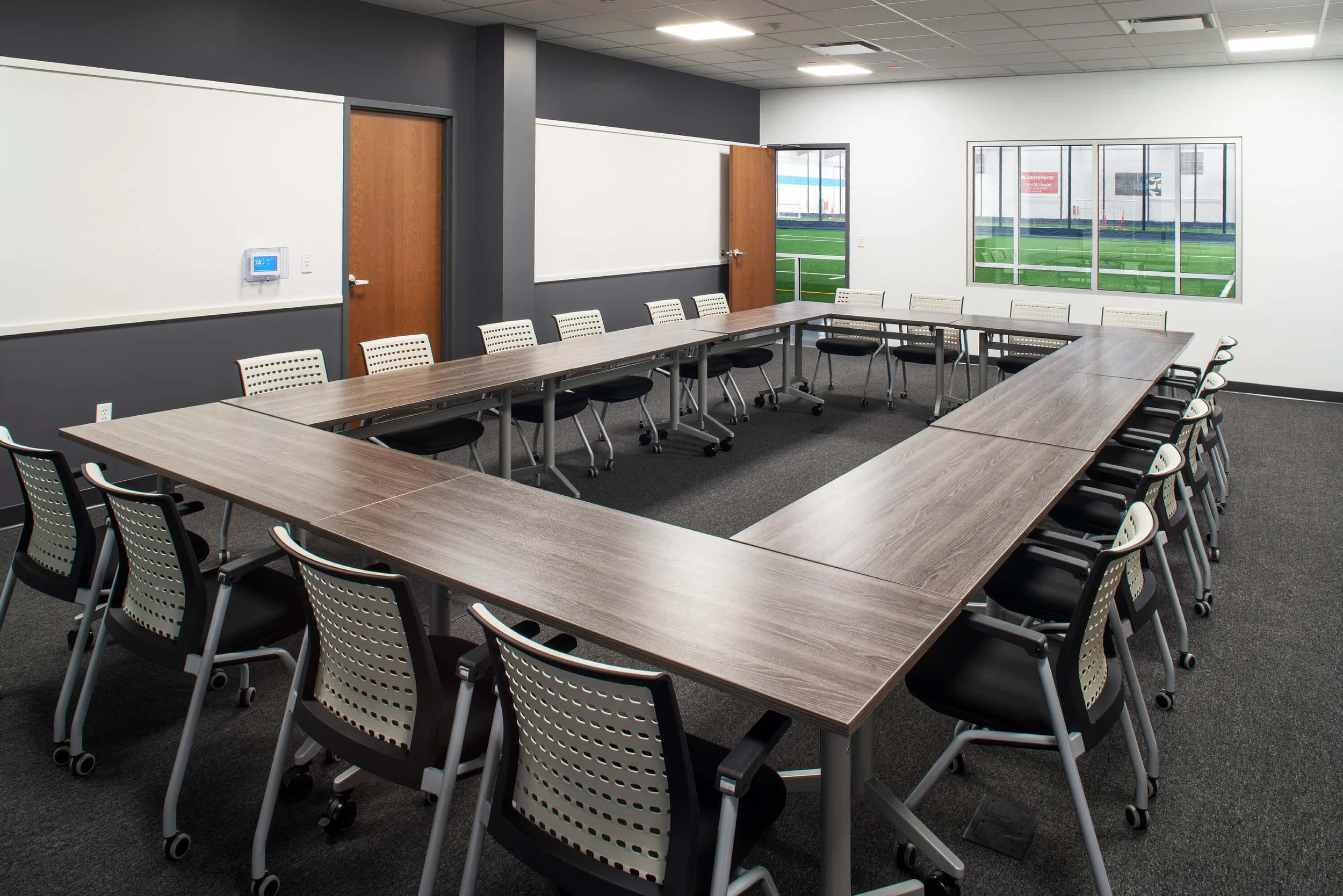 Conference room with U-shaped wooden tables and black and beige chairs, window view of a sports field, white and gray walls, digital thermostat on wall.