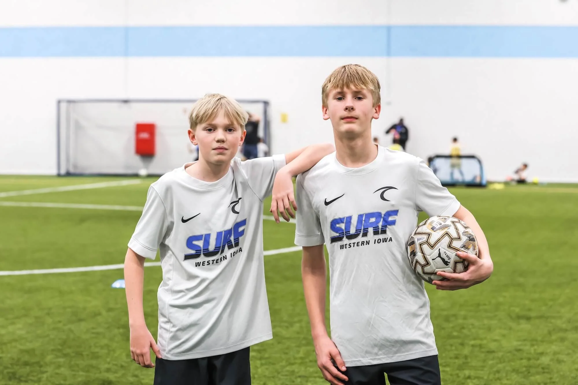 Two young boys in white soccer jerseys with 'SURF Western Iowa' logo, standing on an indoor soccer field. One boy has his arm over the other's shoulder. The boy on the right holds a soccer ball under his arm. In the background, other players can be s