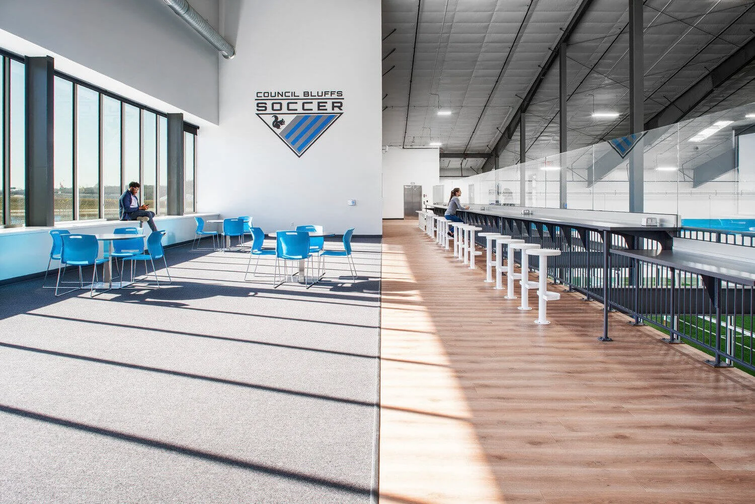 Indoor view of a soccer facility lounge with large windows, blue chairs, tables, and a bar area with stools, at Council Bluffs Soccer.