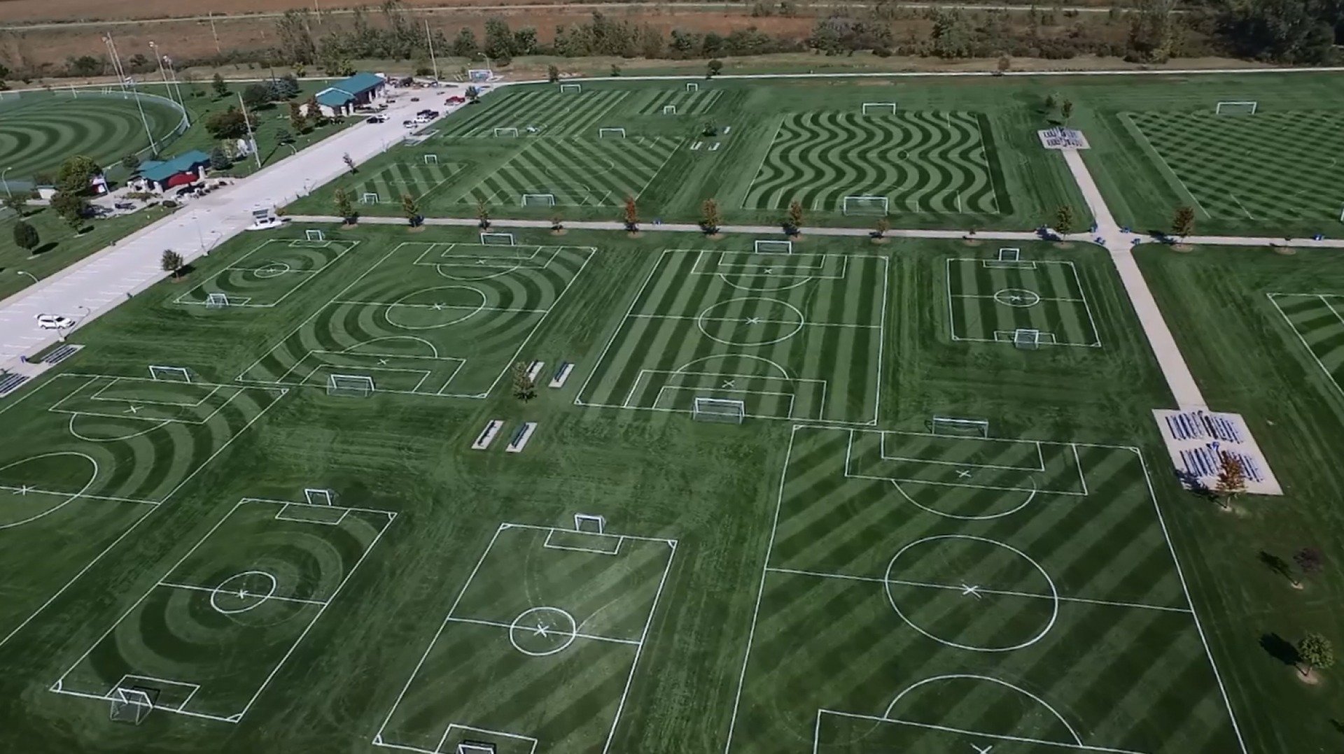 Aerial view of multiple outdoor soccer fields with striped grass and white boundary lines, arranged in a grid pattern, with parking lot, trees, and small buildings around the perimeter.