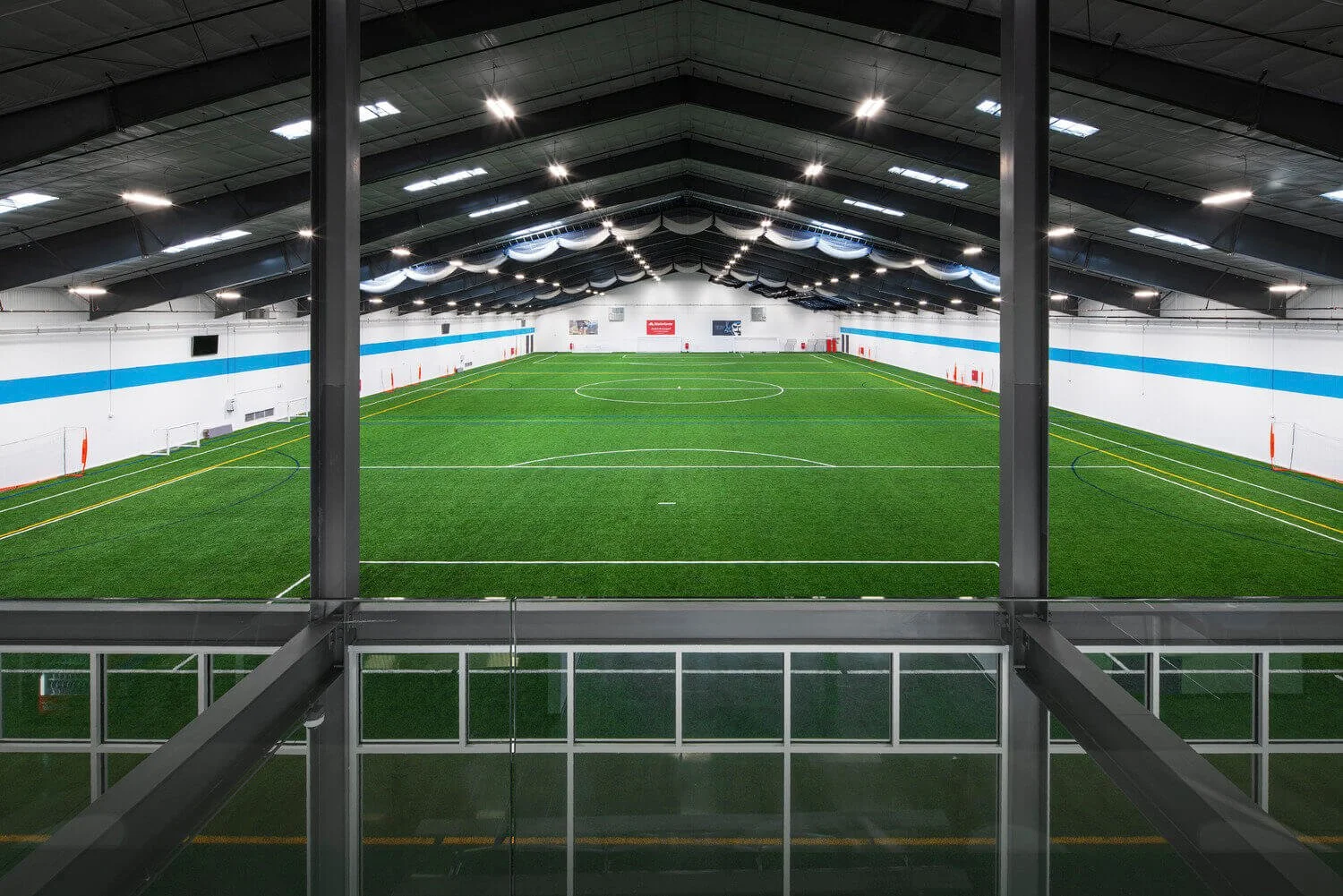 Indoor soccer field with artificial turf, enclosed by white walls with blue stripe, illuminated ceiling lights, goals at each end, and viewing balcony with glass railing in the foreground.