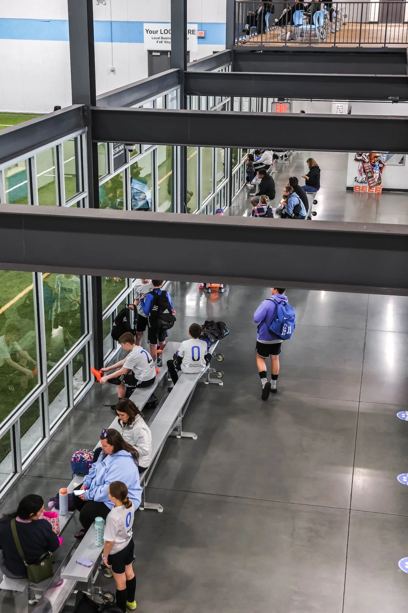People sitting and standing in a sports facility with large windows showing a green field outside, some kids in soccer uniforms, and a few adults waiting, some using their phones.