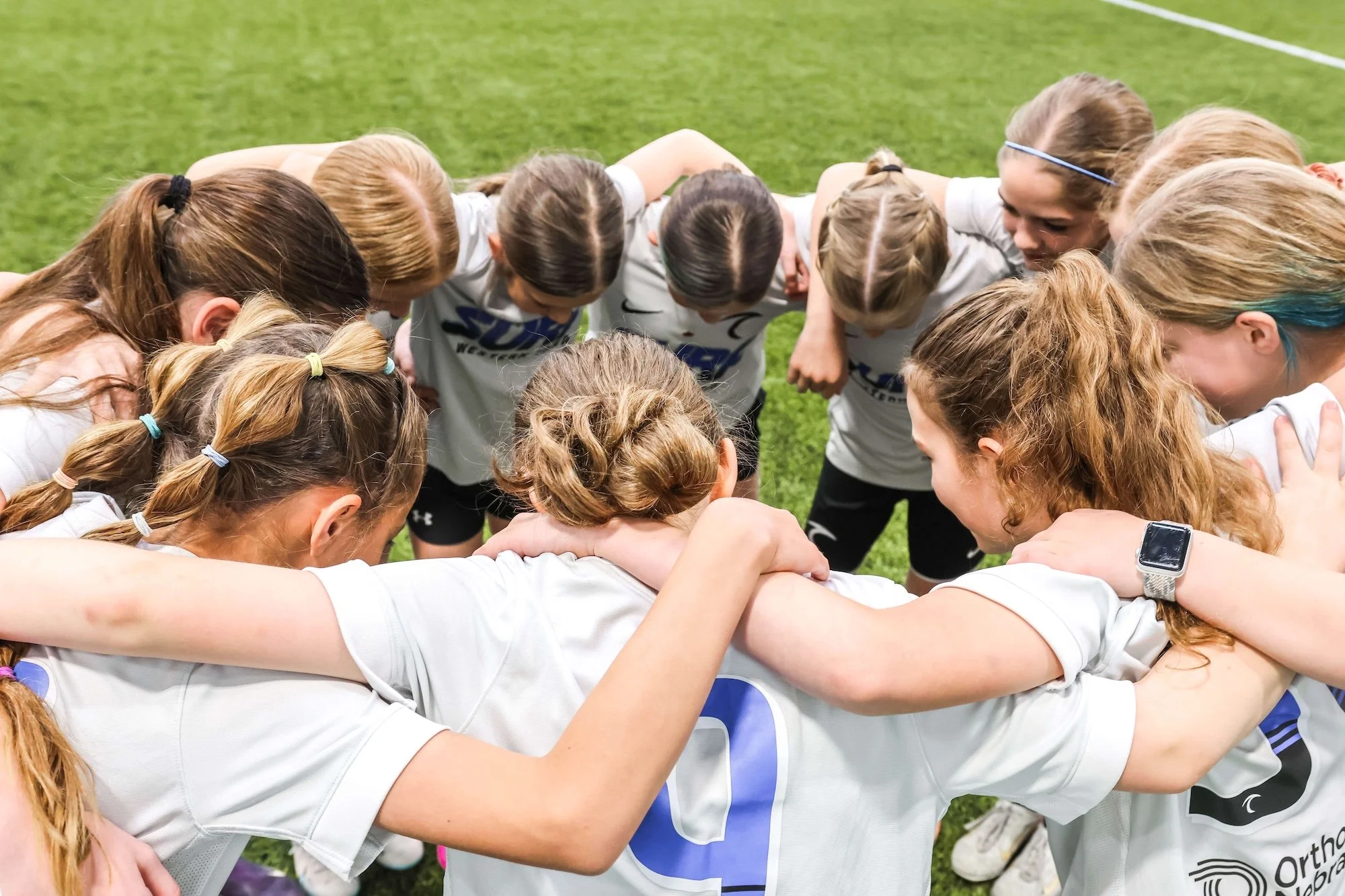 A group of female soccer players huddling together in a circle on a green field, with their arms around each other's shoulders.