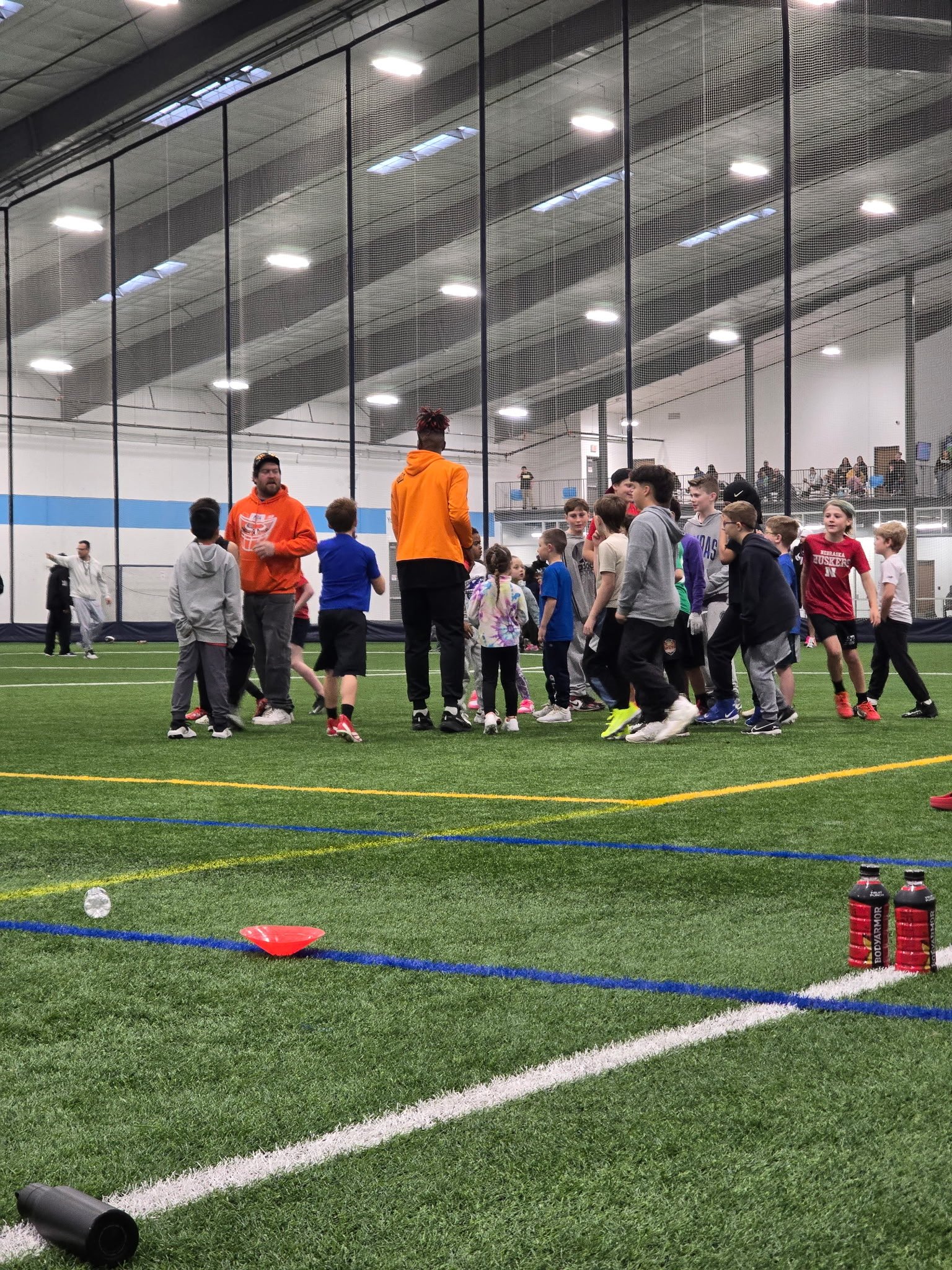 Children and coaches gather on an indoor sports field with artificial turf, inside a large enclosed gym with high ceilings and netting dividing the space, with spectators watching from above.