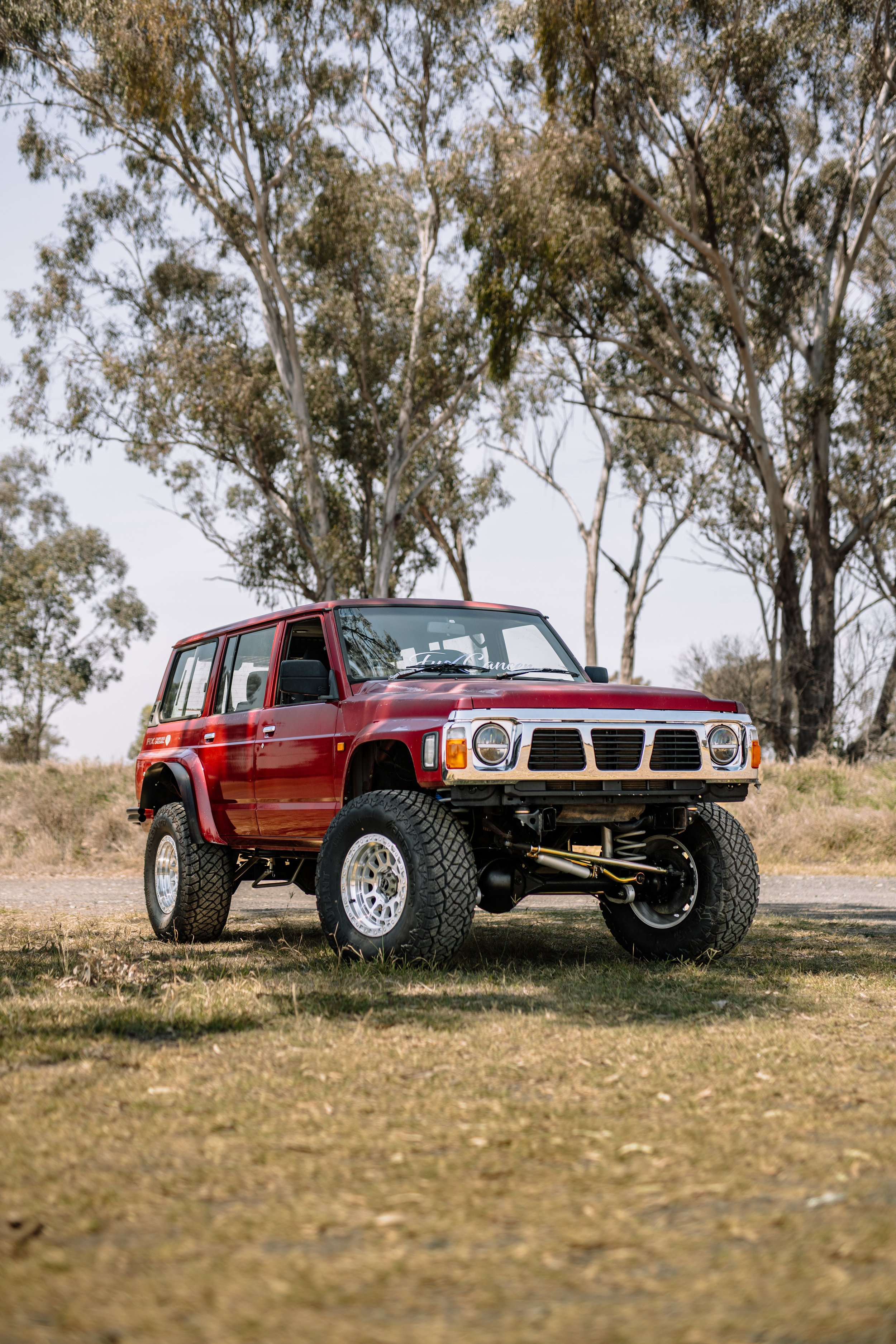 A red lifted off-road vehicle with large tires, parked outdoors on grass with trees in the background.