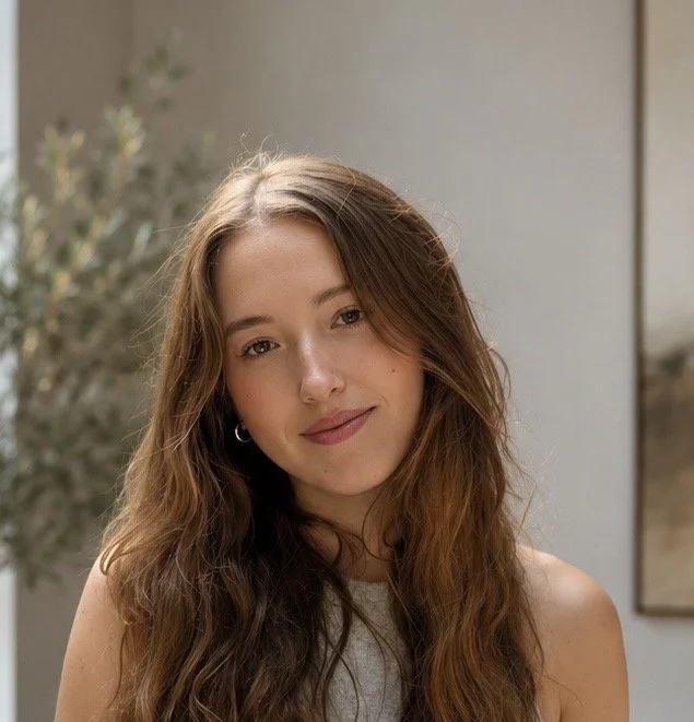 A young woman with long, wavy brown hair and a subtle smile, wearing a sleeveless top, standing indoors with a blurred background and a mirror on the wall.