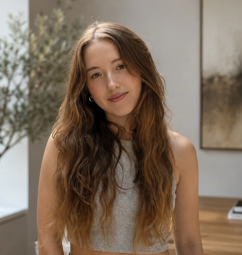 A young woman with long, wavy brown hair wearing a gray sleeveless top, smiling softly in an indoor setting with neutral walls and a plant in the background.