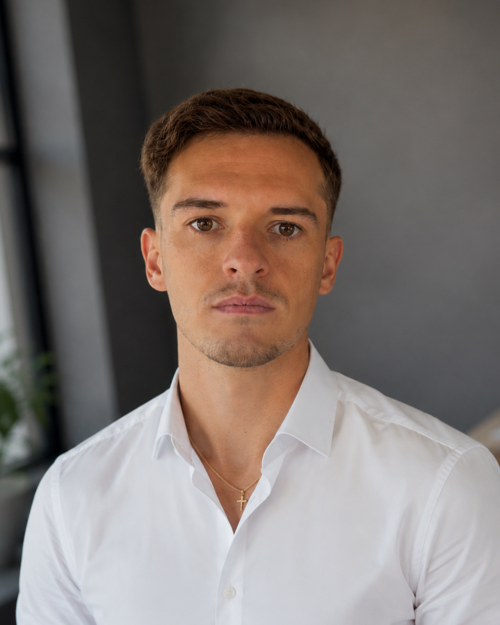 A young man with short brown hair and light skin wearing a white dress shirt and a gold chain with a cross pendant, standing indoors against a dark wall with windows and greenery in the background.