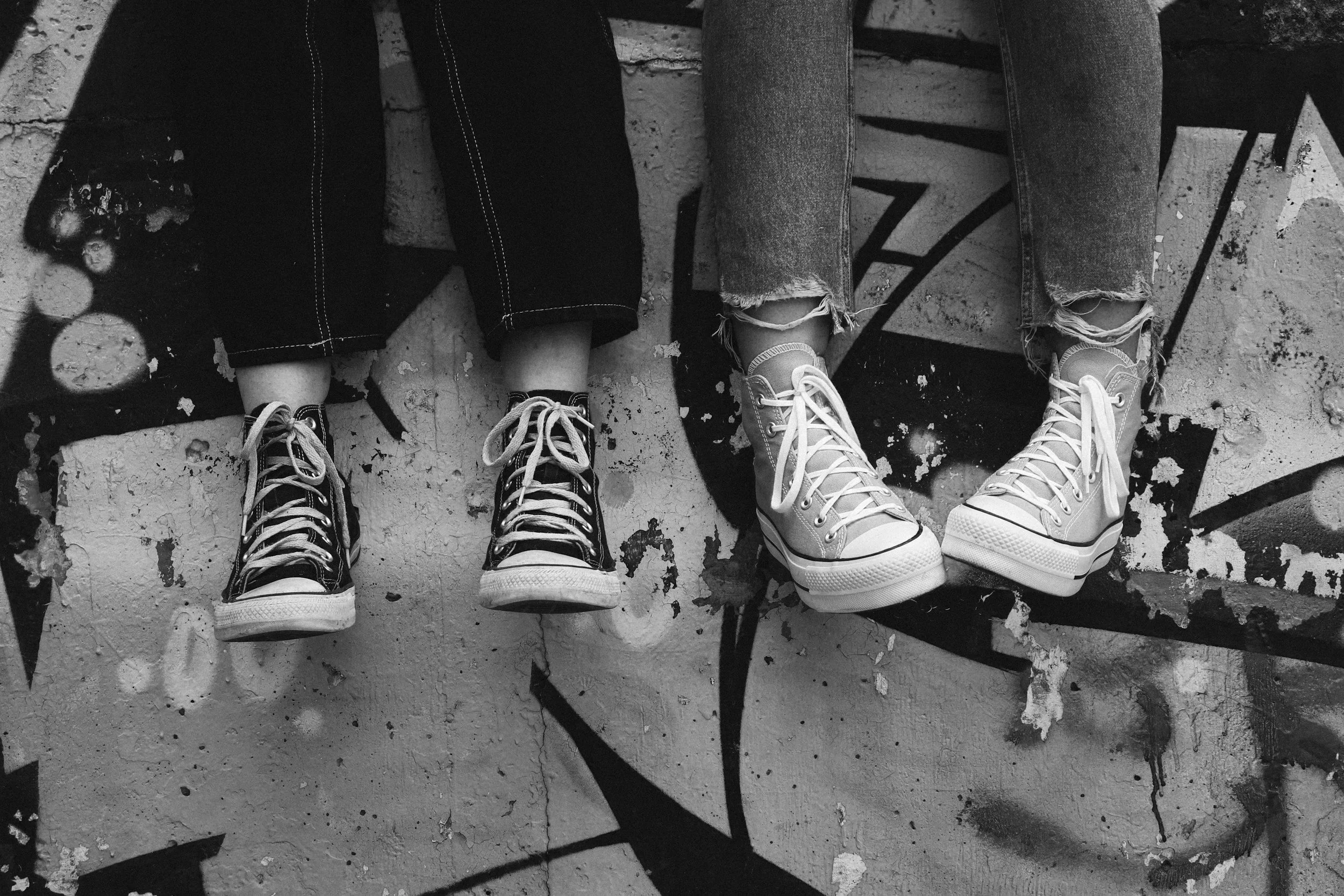 Close-up of two people sitting on a bench with graffiti on the wall behind them, wearing sneakers and ripped jeans.