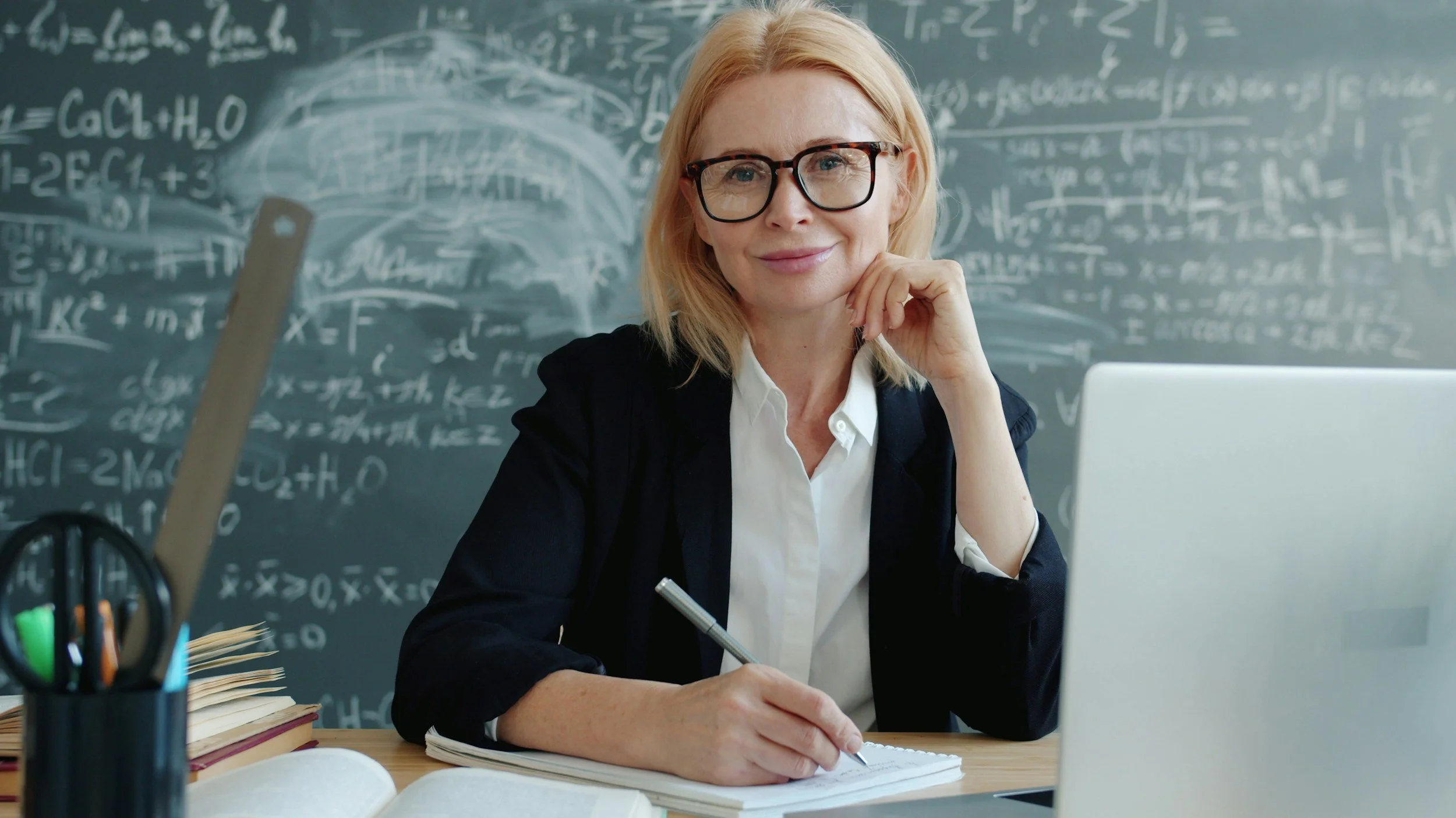 A woman wearing glasses sitting at a desk in front of a chalkboard with mathematical formulas, holding a pen in her right hand and looking at the camera.
