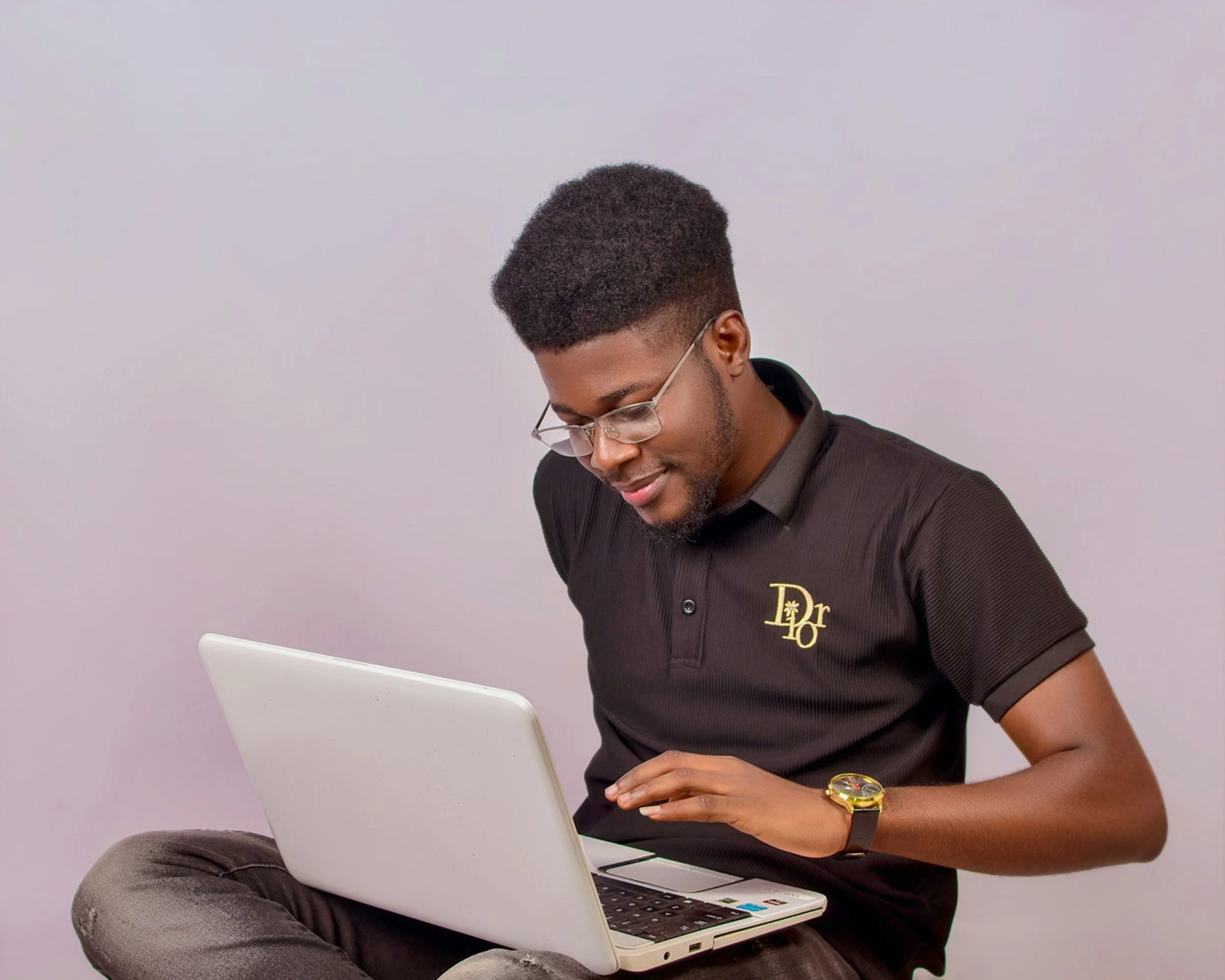 A young man with glasses and a black polo shirt sits cross-legged while using a white laptop.