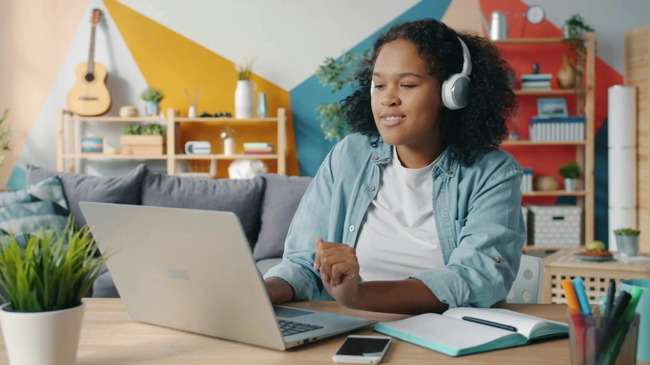 A woman with curly hair wearing headphones and a denim shirt, sitting at a desk with a laptop, in a colorful modern living room.