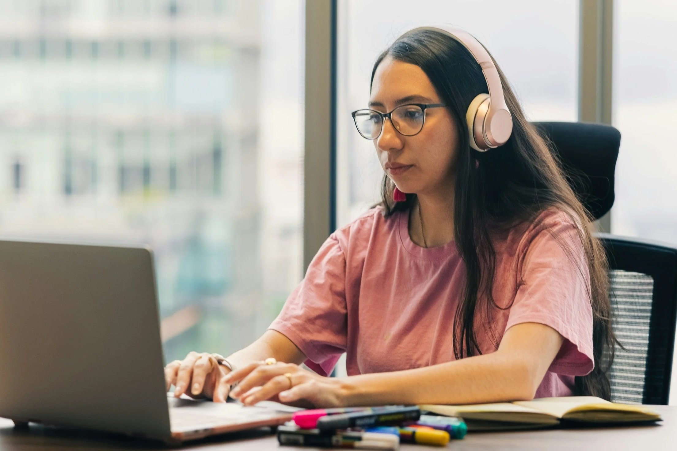 Young woman with glasses and long dark hair working on a laptop at a desk, wearing large pink headphones, with markers and an open notebook nearby, in an office with large windows.