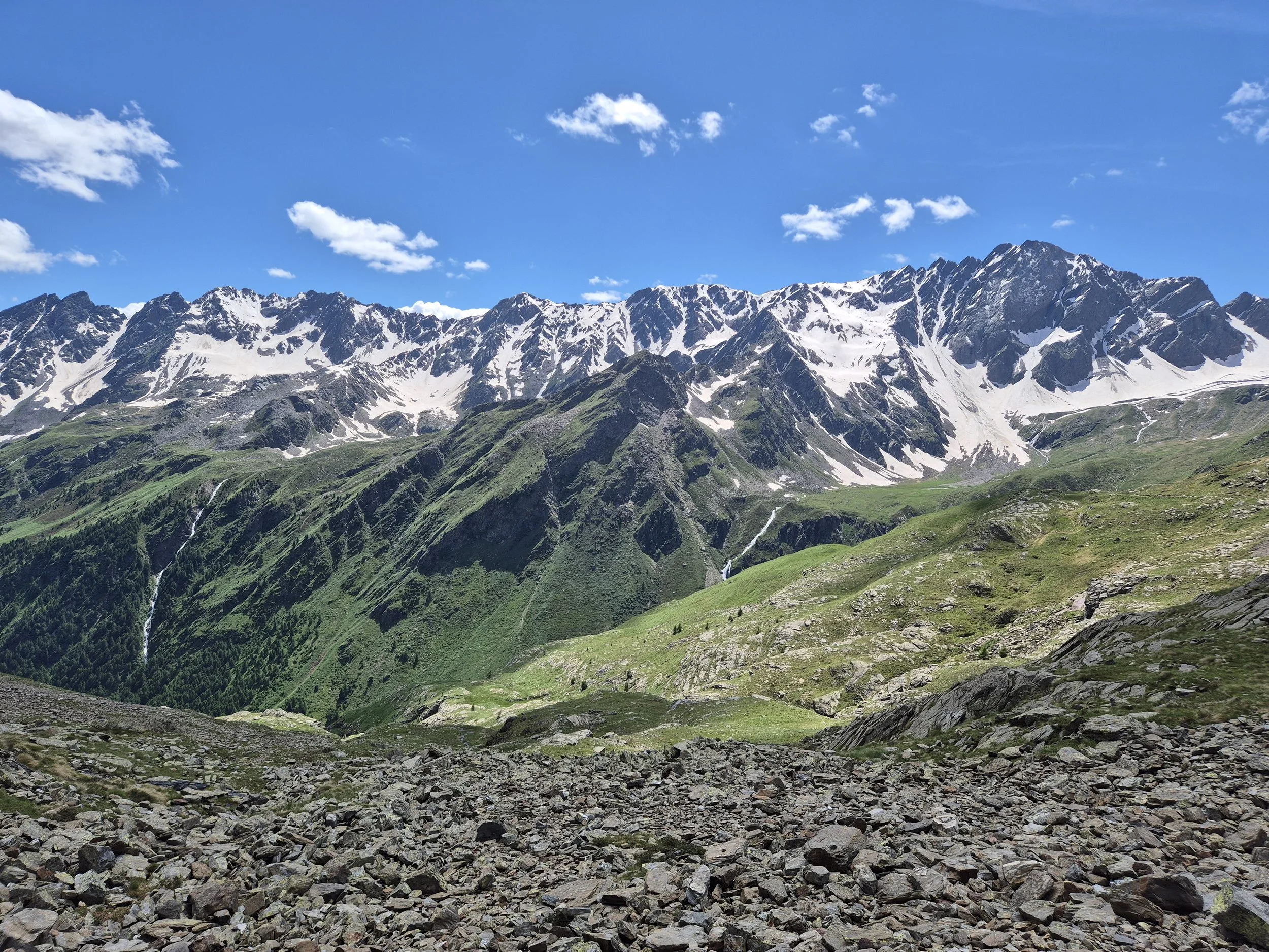 Mountain landscape with snow-capped peaks, green valleys, and rocky foreground under a blue sky with a few clouds.