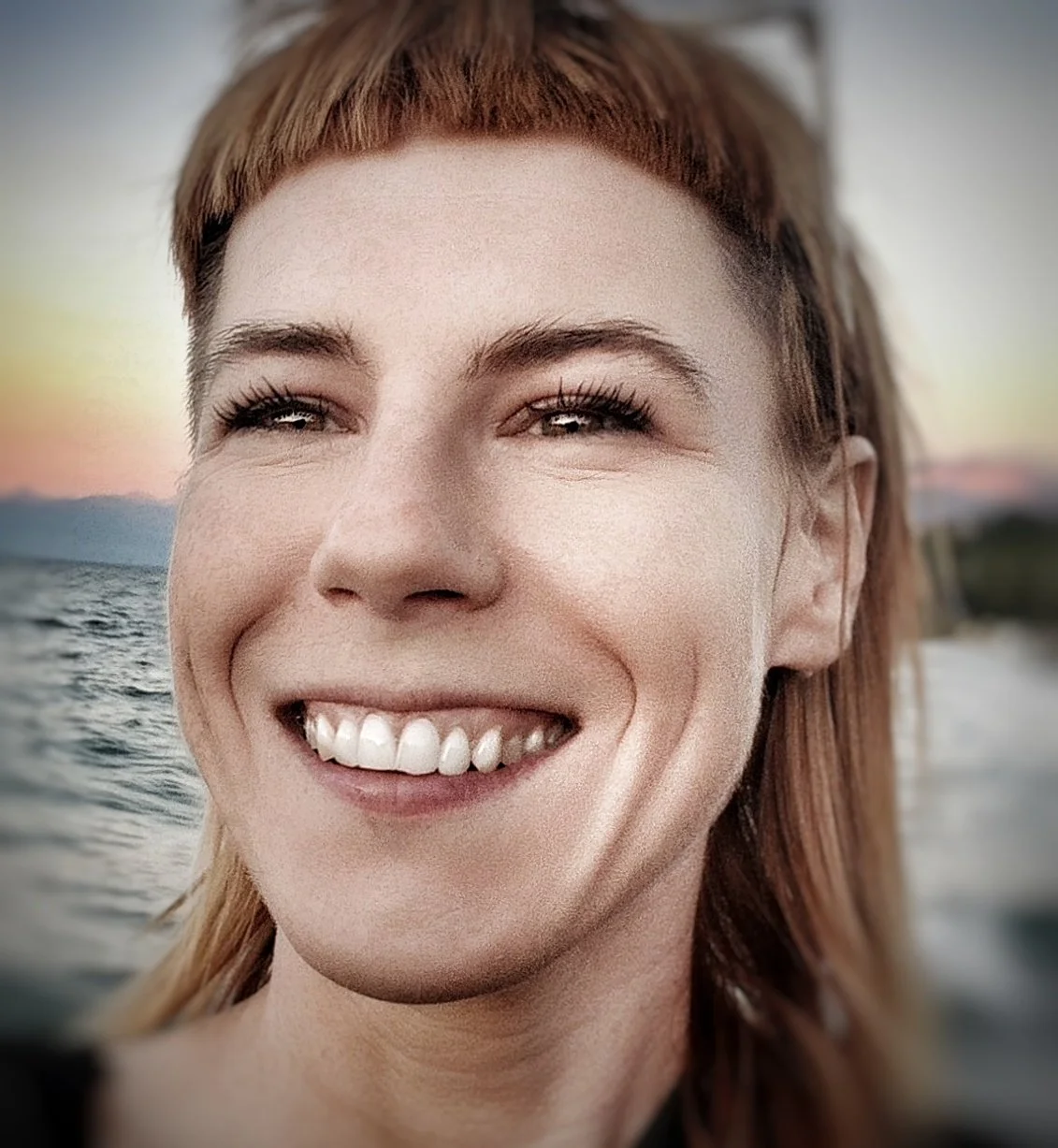 Close-up of a woman with red hair smiling, with a body of water and sunset in the background.