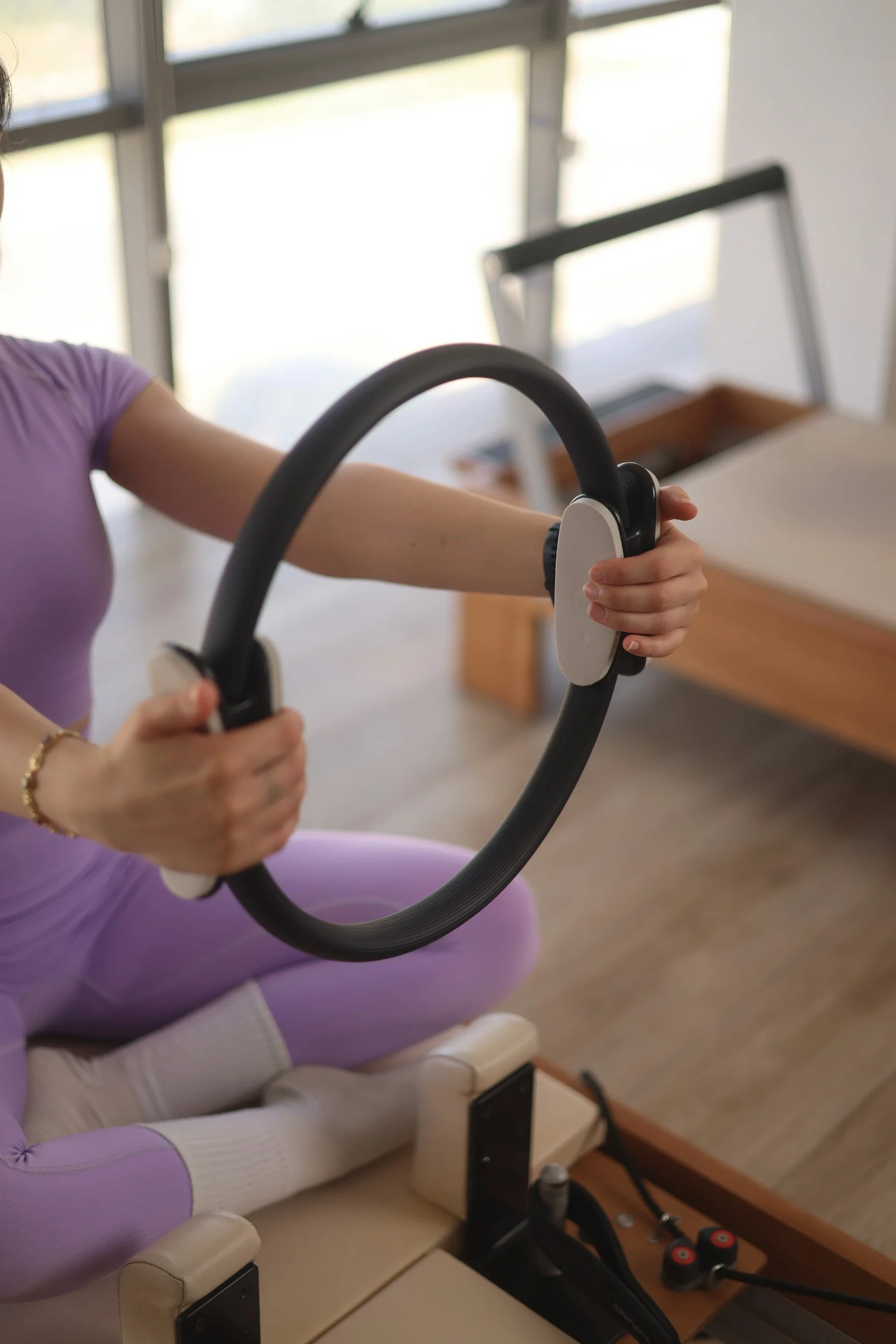 Person wearing purple leggings and a purple top exercising on a Pilates reformer machine in a well-lit room with large windows.