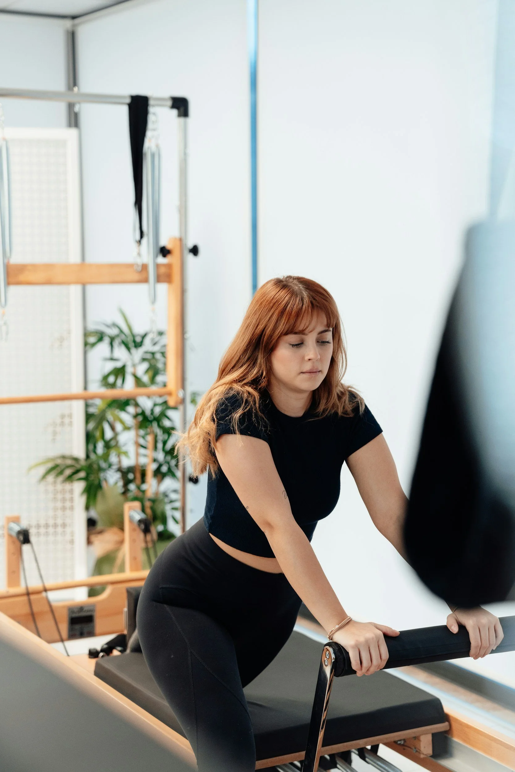 A woman with wavy auburn hair stretching on a pilates reformer machine in a fitness studio, with a white wall background and gym equipment and plants in the room.