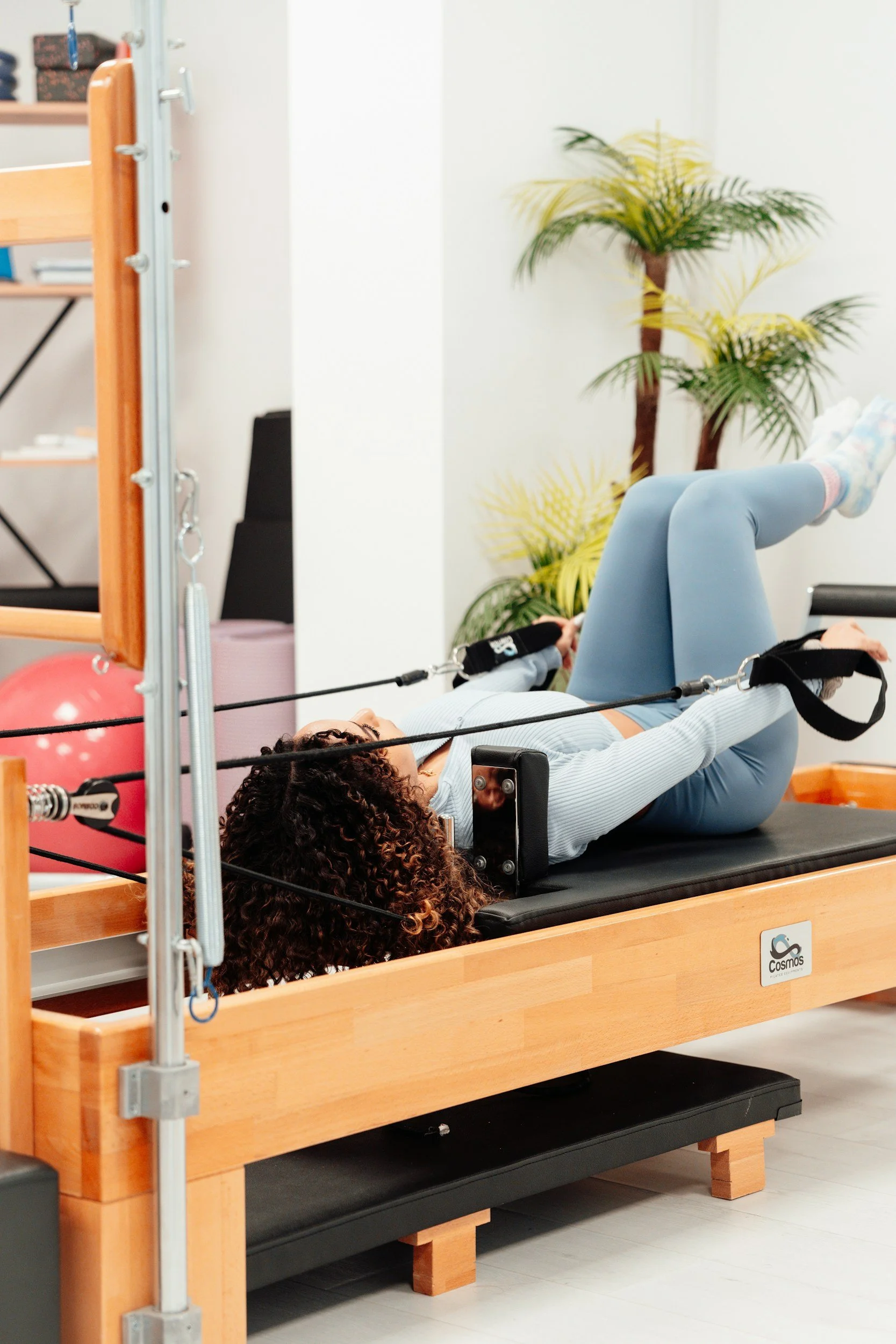 A person lying on a Pilates reformer machine during exercise, with their legs raised and bent at the knees. The setting is a fitness studio with a wooden reformer and a background decorated with indoor plants and a mirror. The person has curly hair and is wearing a light-colored top and blue leggings.
