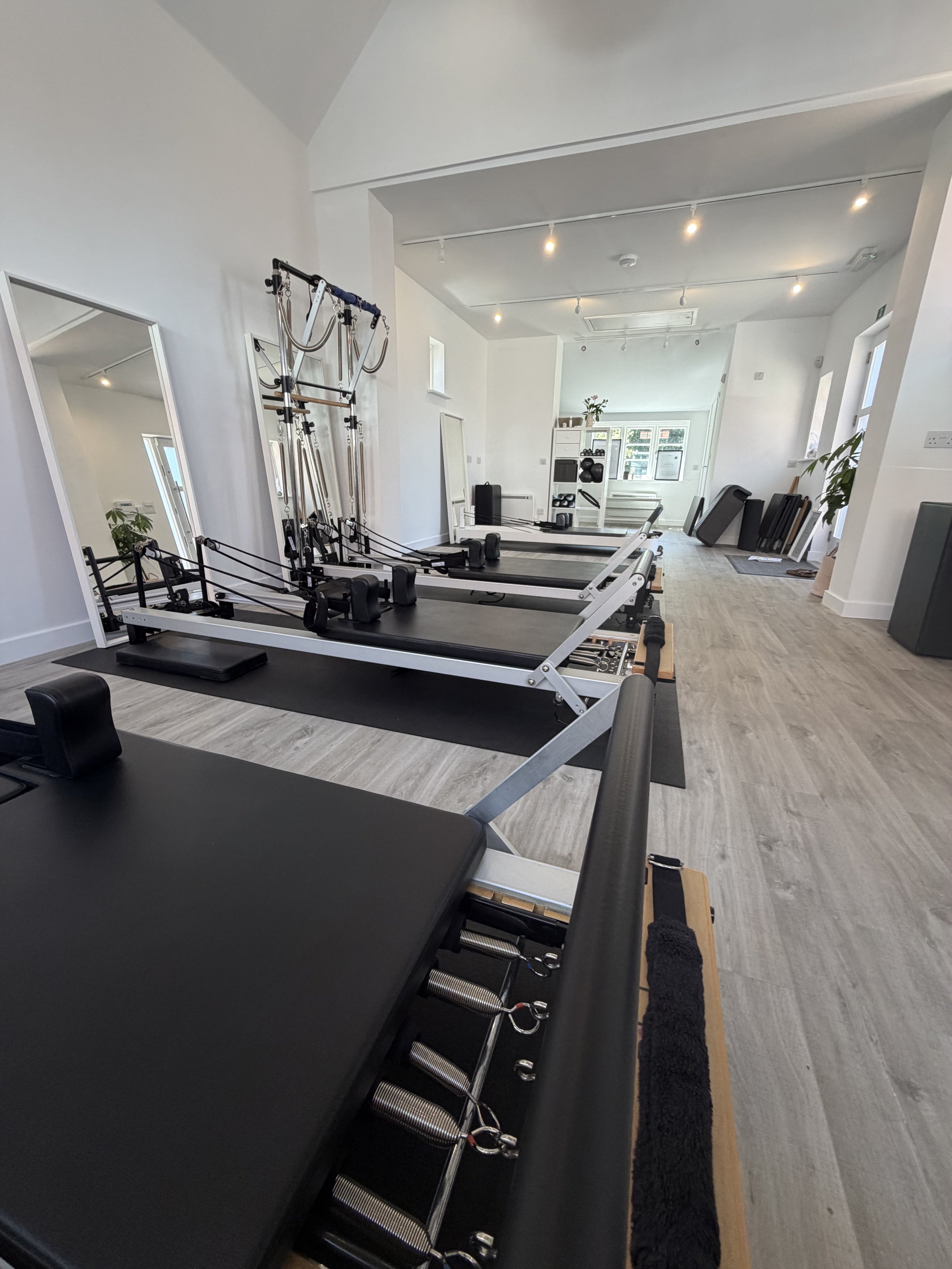Pilates studio in Grayshott with Pilates reformer machines lined up, large mirror, wooden flooring, white walls, and exercise equipment in the background.